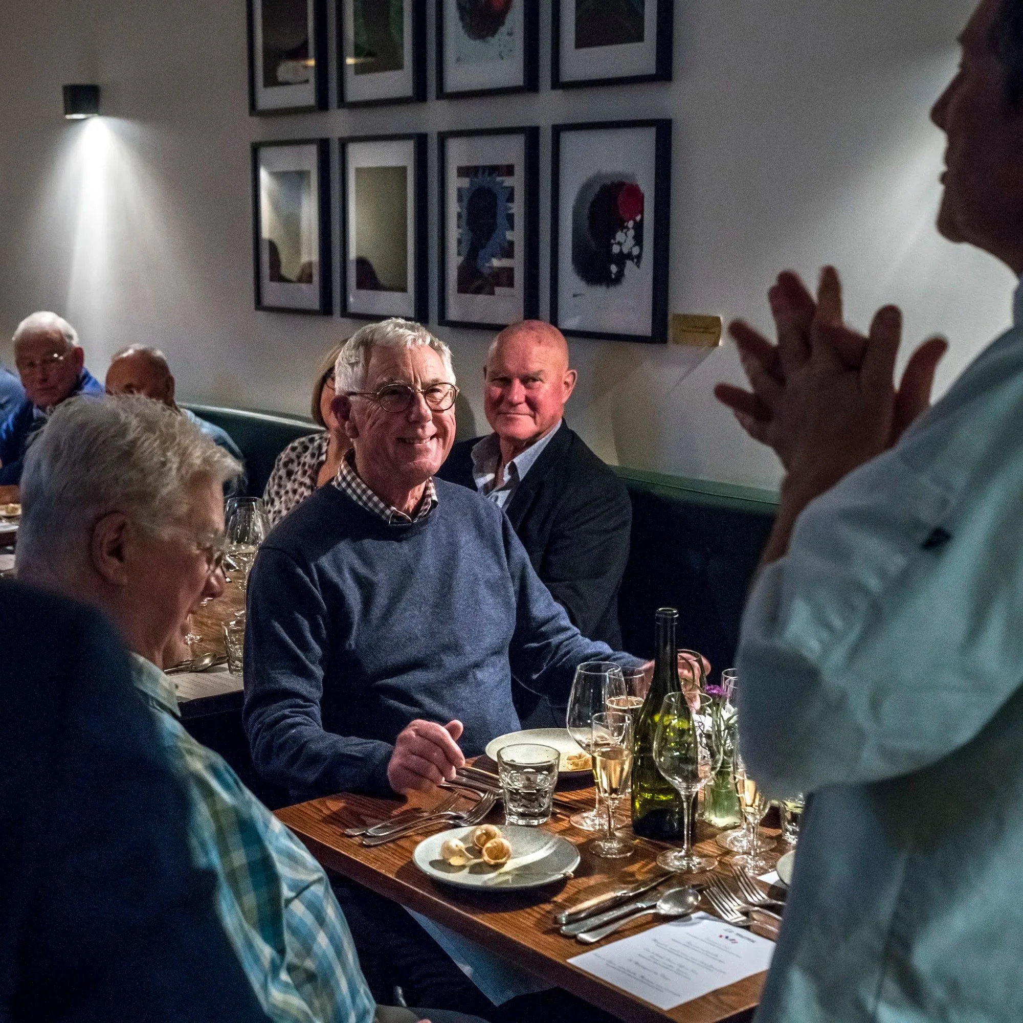 A group of people sitting at a dining table in a dimly lit restaurant, engaging in conversation. The table is set with glasses, cutlery, and a small plate of waffles. The background features a wall decorated with framed pictures.