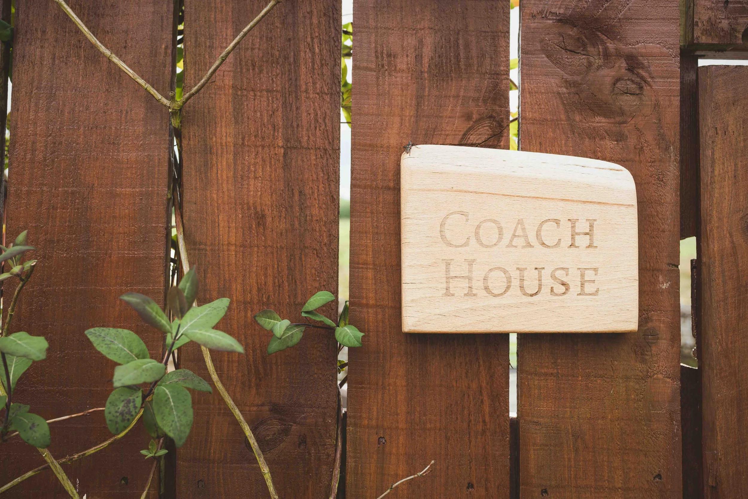 A wooden sign engraved with the words 'Coach House' hanging on a wooden fence with greenery around.