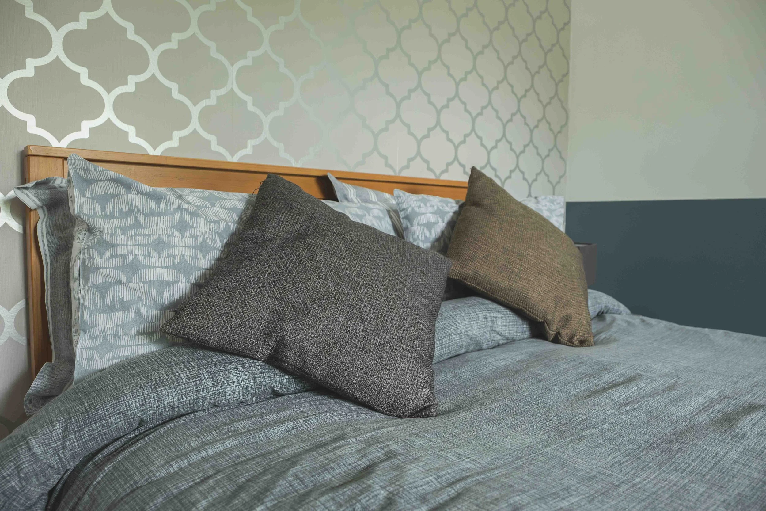 A neatly made bed with a wooden headboard, decorated with several pillows in shades of gray and brown, set against a wall with a geometric patterned wallpaper.