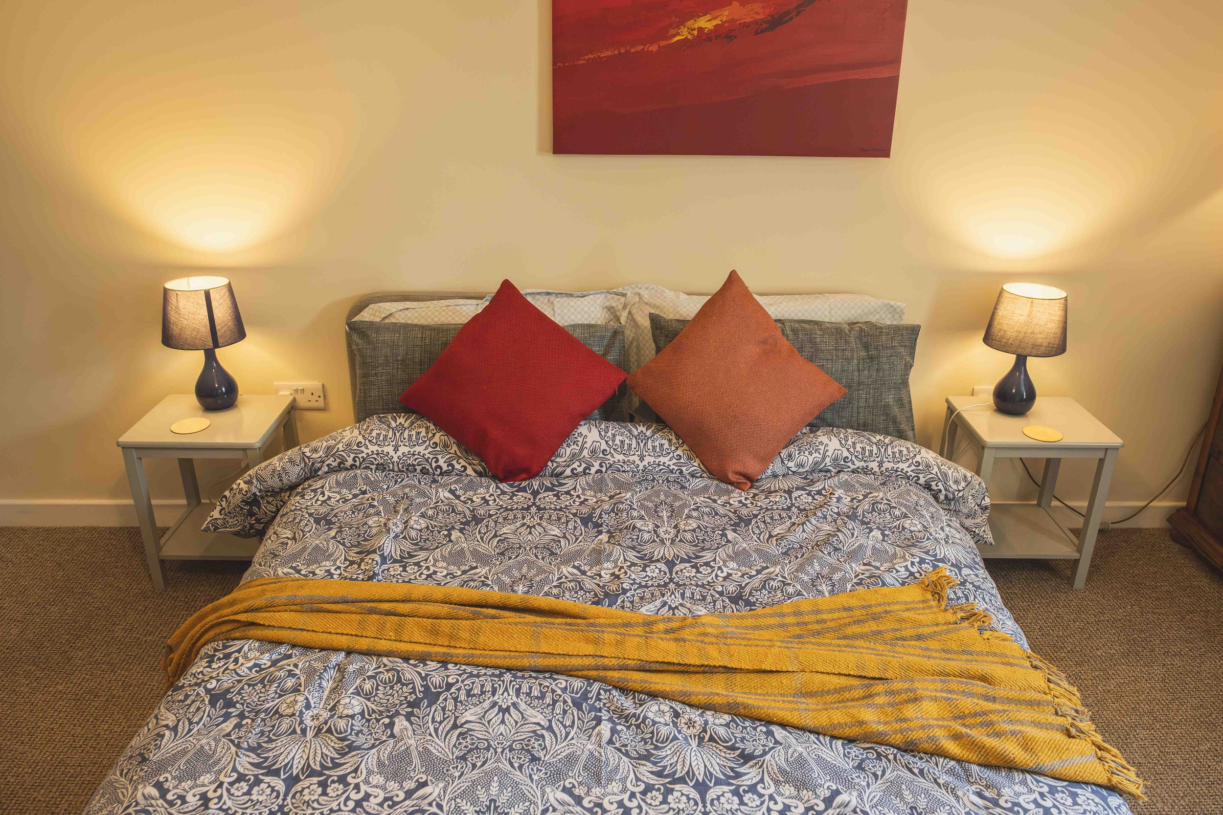 A bedroom with a bed featuring a paisley-patterned bedspread, two red pillows, and two gray pillows. There are two white nightstands with lamps on each side of the bed, and a large red abstract painting hanging above the headboard.