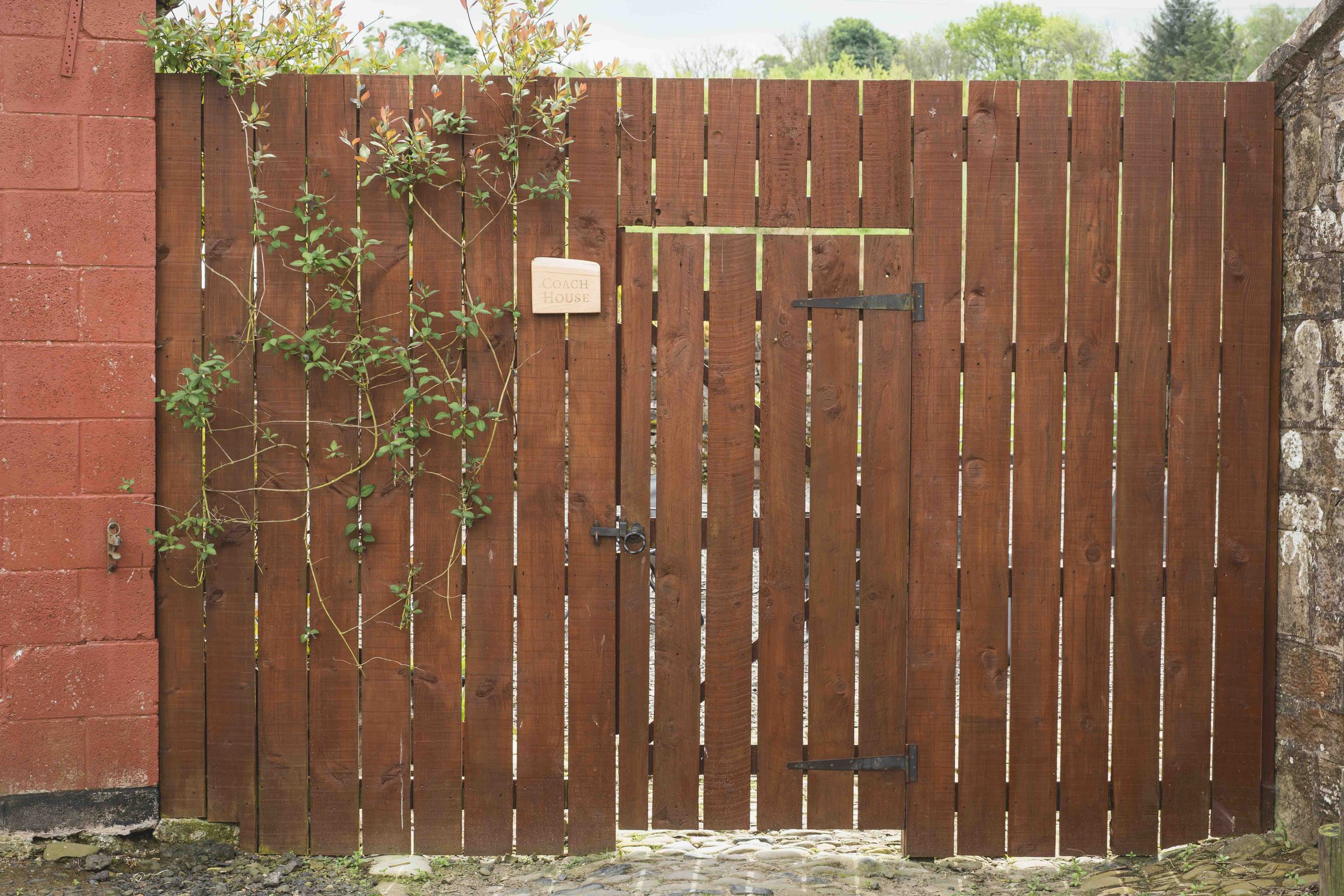 A wooden gate with vertical planks, mounted between a brick wall on the left and a stone wall on the right, with a small sign reading 'Coach House' attached to the gate. There is a vine growing along the left side of the gate.