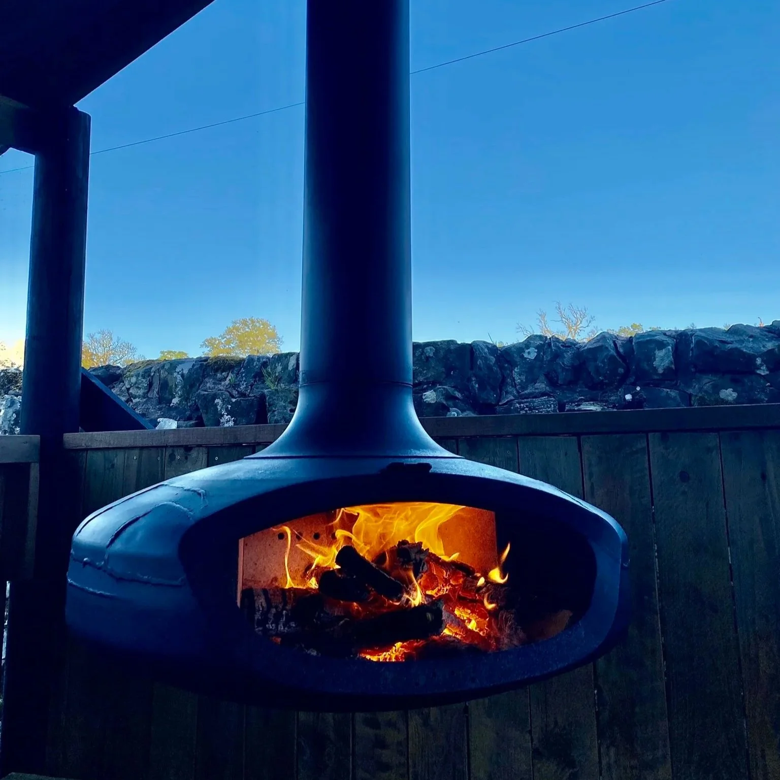 Outdoor hanging fire pit with flames and burning logs, surrounded by a wooden fence and a stone wall, under a clear blue sky.