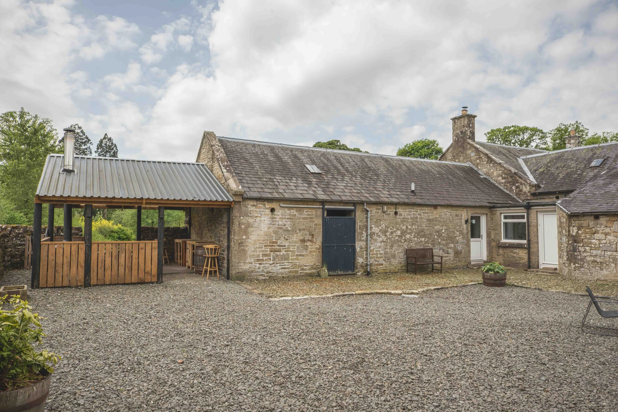 A rustic stone house with a sloped roof, chimney, and a small shed with a metal roof, set in a gravel courtyard with trees and a cloudy sky in the background.