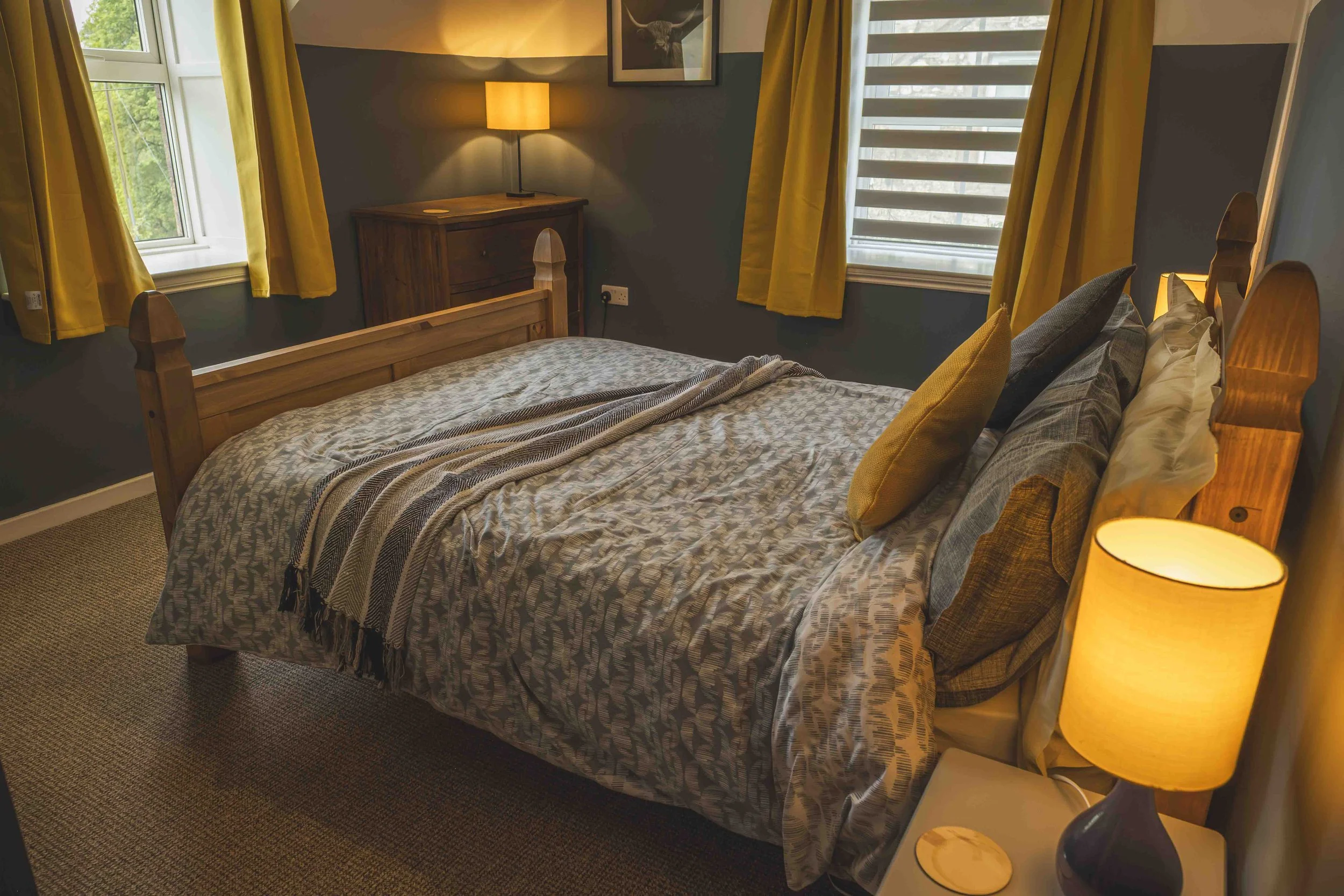 A cozy bedroom with a wooden bed frame, patterned bedspread, multiple pillows, and a nightstand with a lamp. Two windows with yellow curtains allow natural light, and a framed picture of a highland cow hangs on the wall.