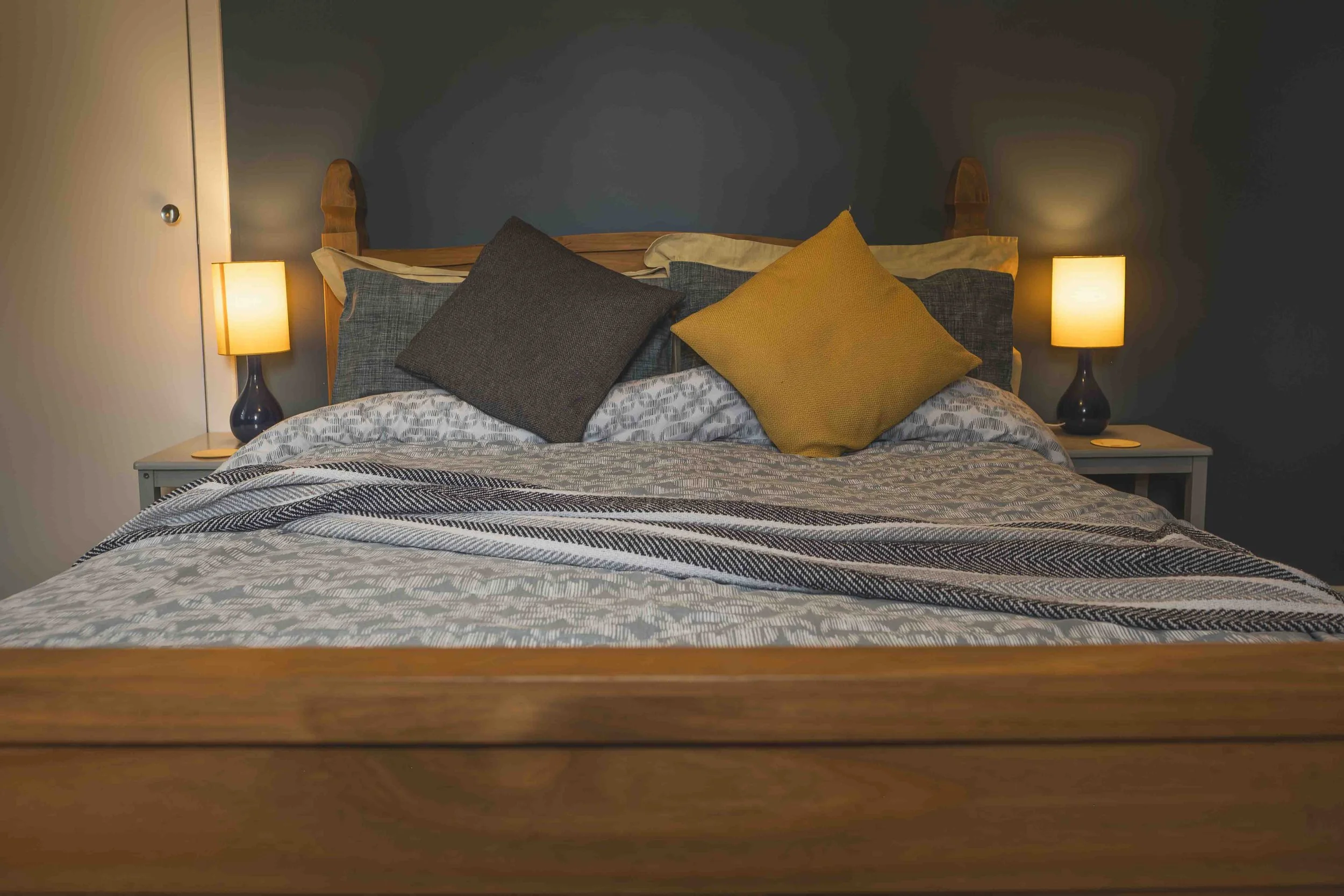 A neatly made bed with multiple pillows and two bedside lamps turned on in a dimly lit room.
