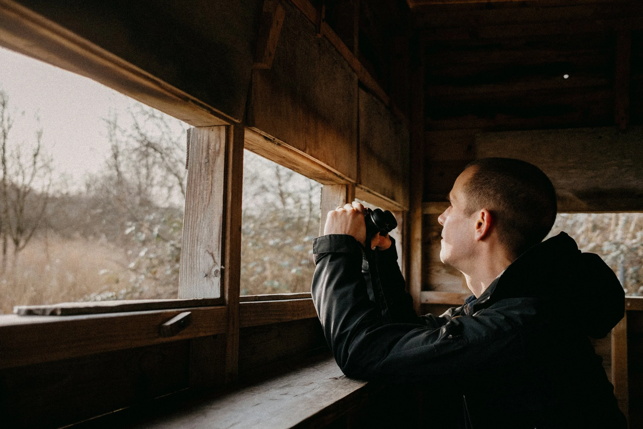 A young man looking through binoculars while sitting inside a wooden structure with open windows, possibly a hunting blind, overlooking a winter landscape with leafless trees.