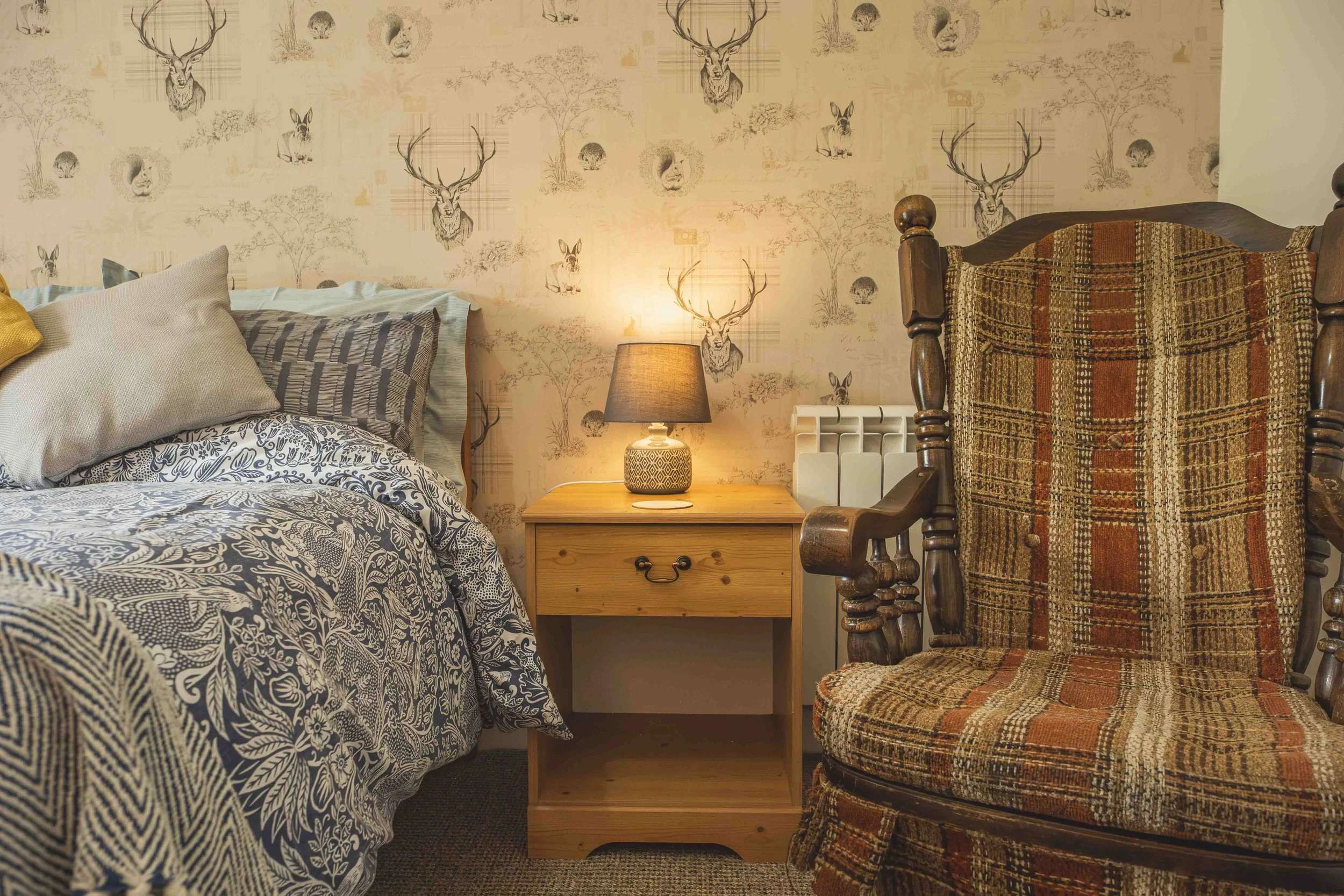 A bedroom with a bed covered in patterned bedding, a wooden nightstand with a lamp, and a vintage wooden chair with a plaid upholstered seat and back near the bed. The wall behind features wallpaper with deer, fox, rabbit, and tree illustrations.