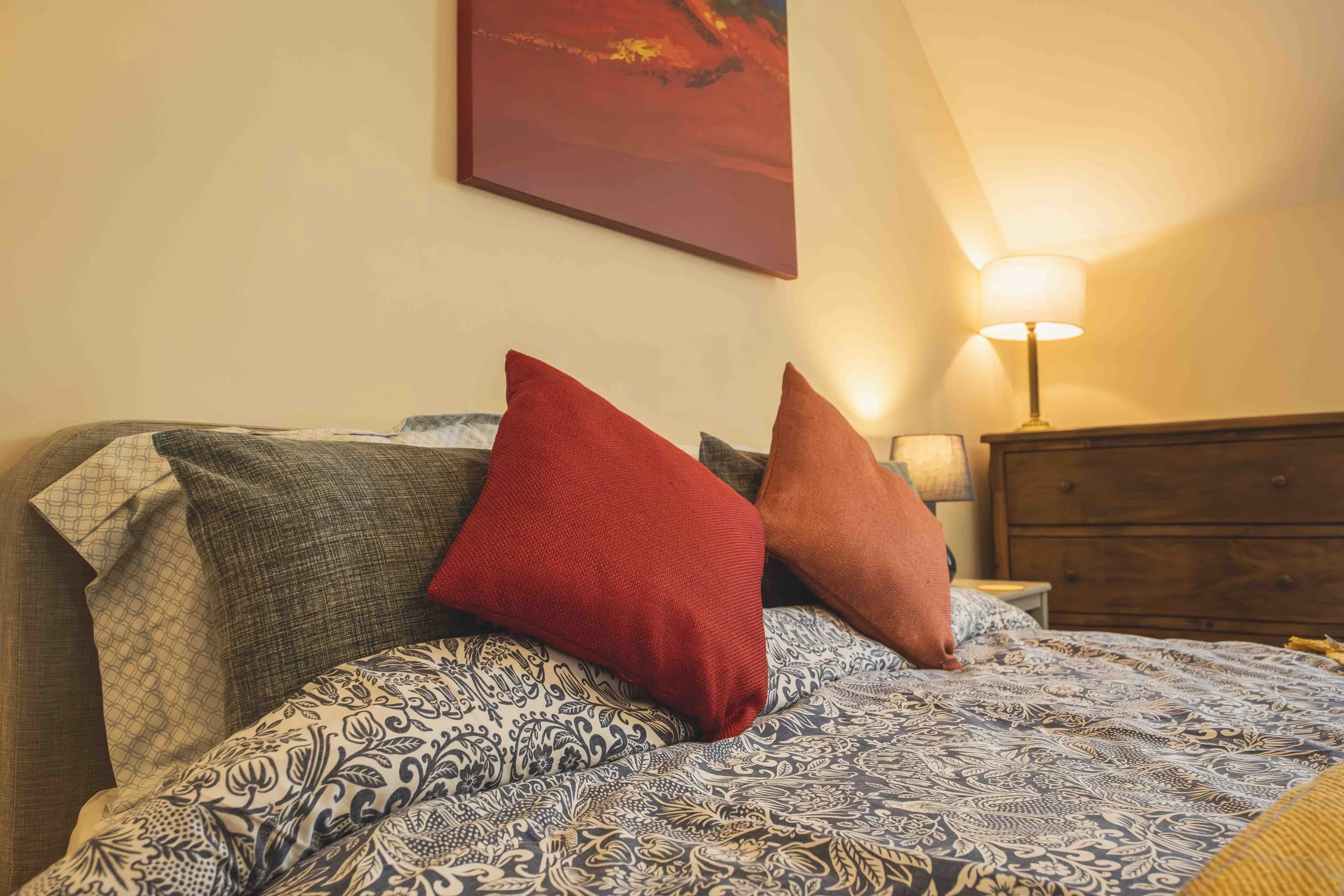 A cozy bedroom with a bed covered in a patterned navy and white bedspread. There are multiple pillows in shades of red, brown, and gray. A wooden nightstand with a lamp is visible, and a red and orange landscape painting hangs on the beige wall.