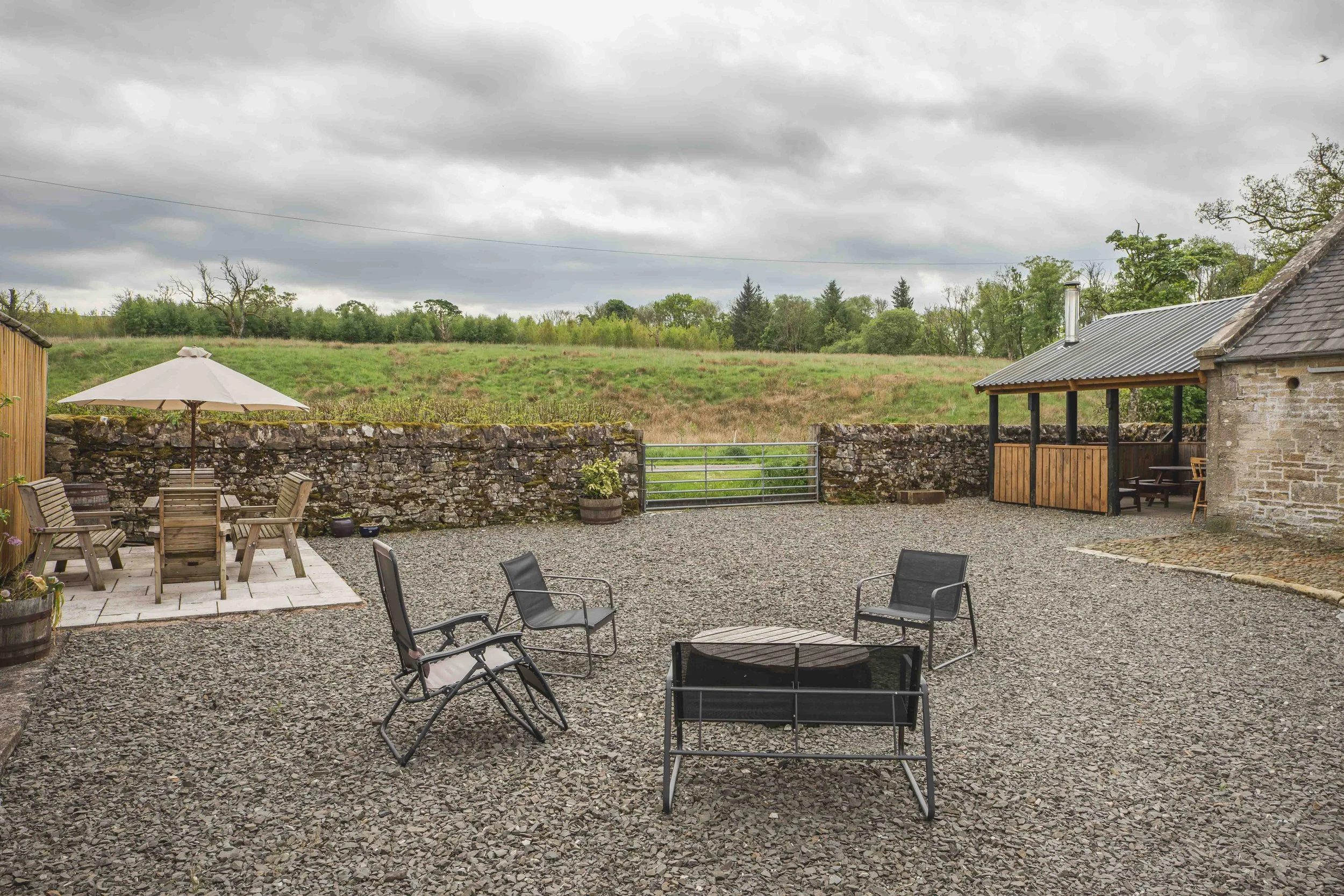 Outdoor patio area with gravel ground, black chairs, a wooden dining table with an umbrella, and a stone building with a metal roof, surrounded by a stone wall and a scenic green hill with trees in the background.