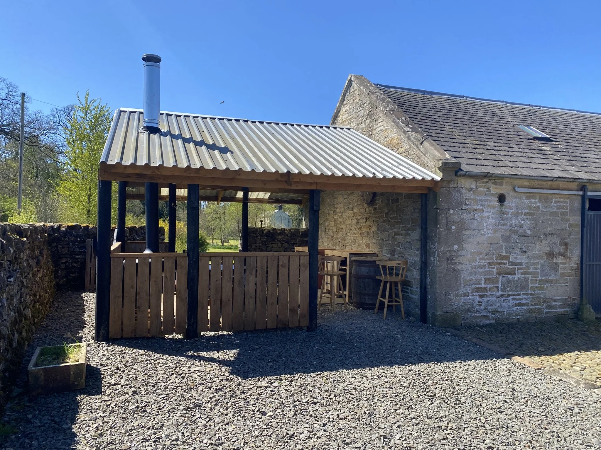 A stone and brick house with a sloped shingle roof and a chimney. In front, there's an undercover barbecue room with a corrugated metal roof, supported by black posts. The ground is covered with gravel, and the background has green trees.
