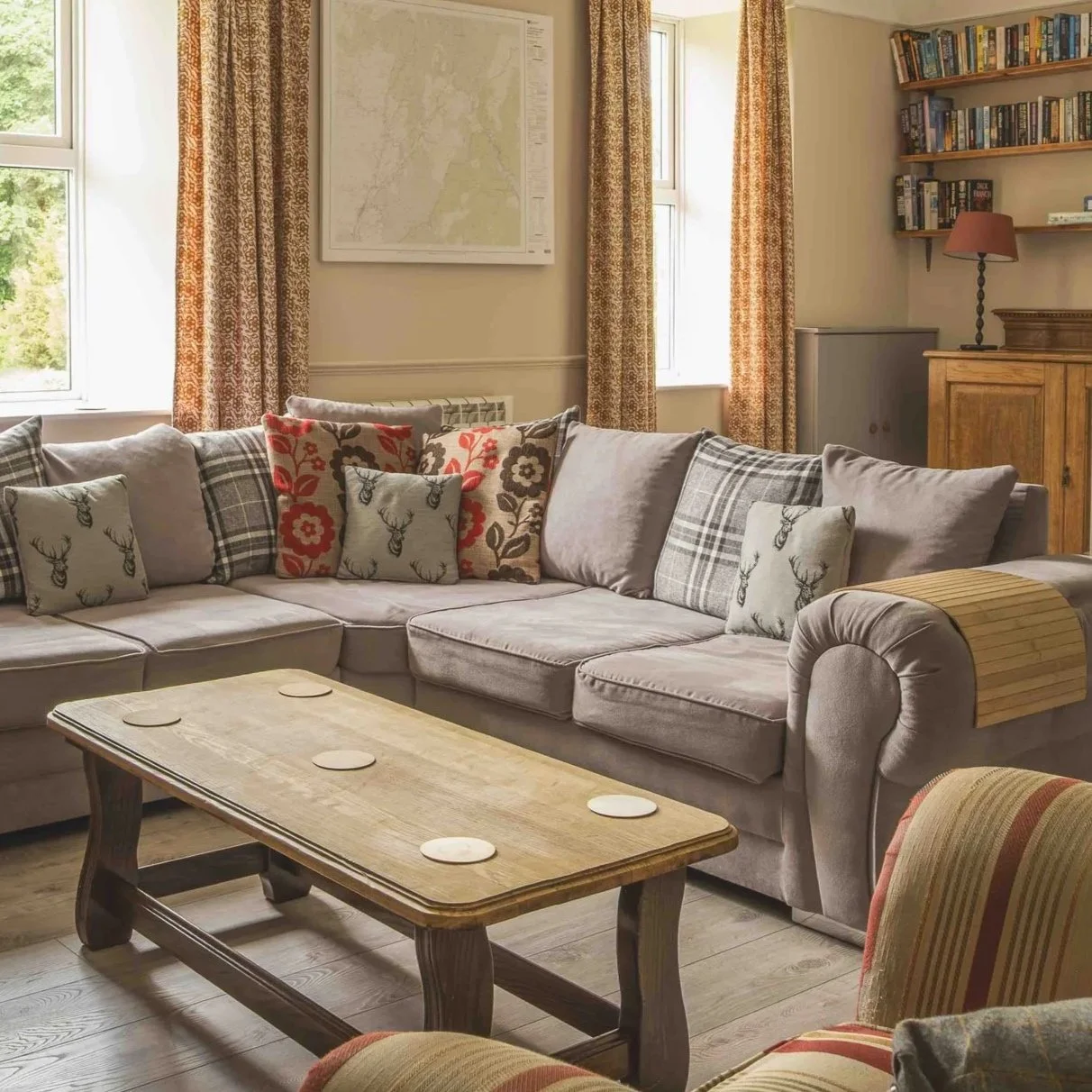 A cozy living room with a beige L-shaped sofa decorated with patterned cushions, a wooden coffee table, large windows with patterned curtains, a wooden bookshelf, and a wooden cabinet with a lamp.
