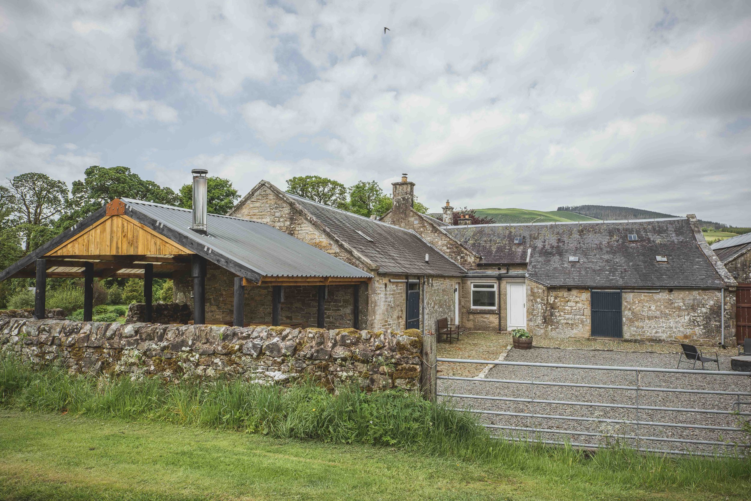 View of a rustic stone house with a metal roof, located in a rural area with green hills in the background. The house has a small courtyard enclosed by a stone wall and a metal gate. There is a grassy lawn in the foreground and a cloudy sky overhead.