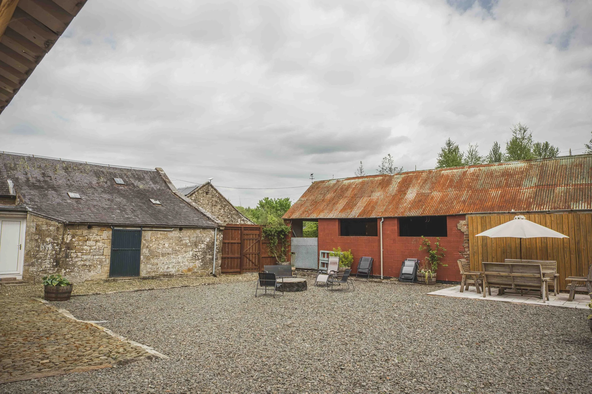 A rustic outdoor patio area with gravel ground, surrounded by stone and wooden buildings, some with tin roofs, and patio furniture including a wooden table and chairs, with an umbrella and green plants. Overcast sky with gray clouds.