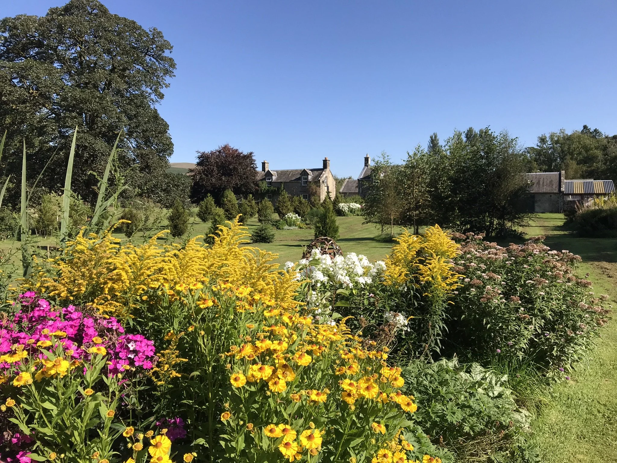 A garden with colorful flowers in the foreground, a house with stone walls and chimneys in the middle ground, and trees and a clear blue sky in the background.