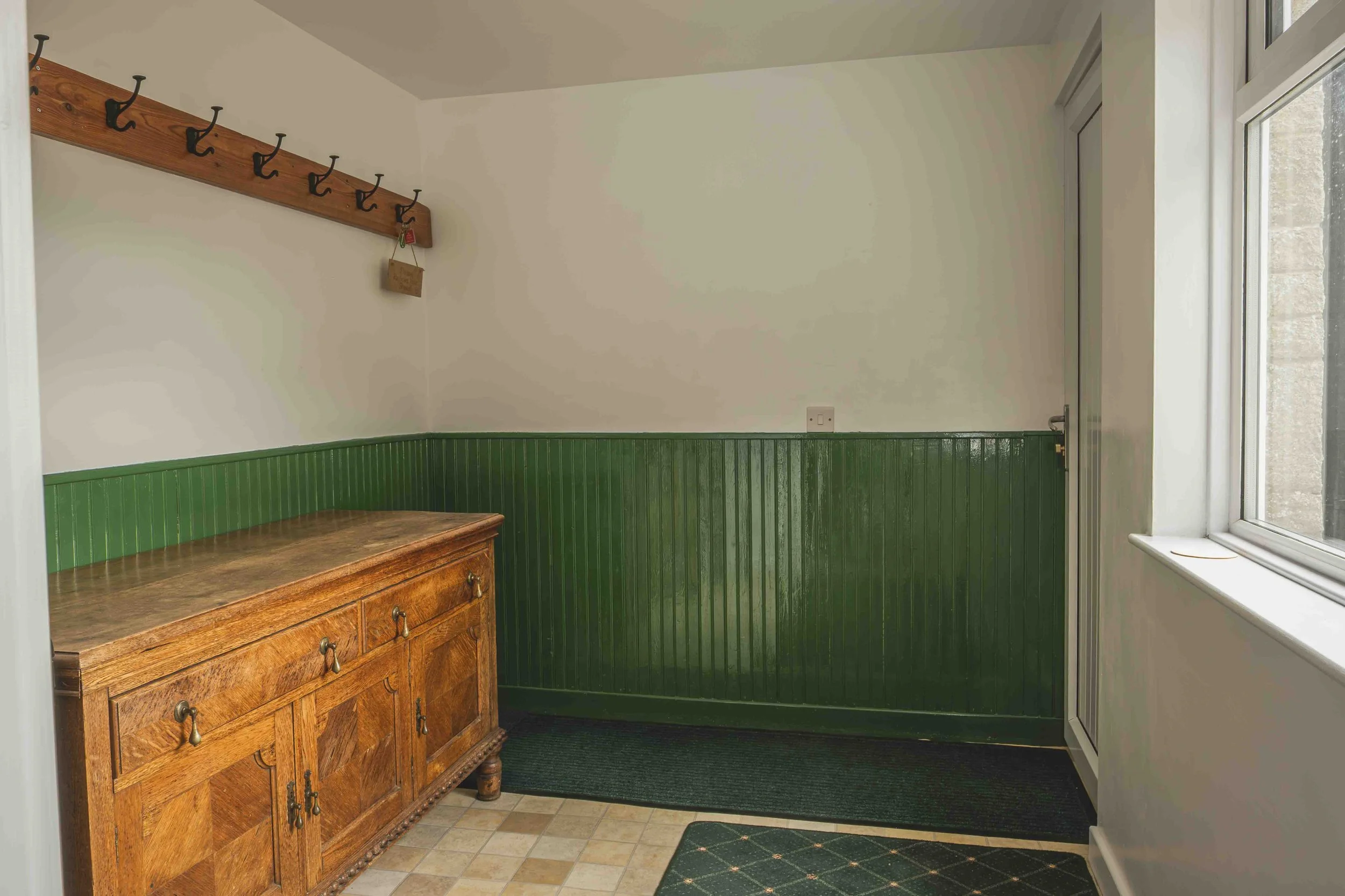 A small entryway with wooden and green paneling walls, a wooden cabinet, a window, and a wooden coat rack with hooks.