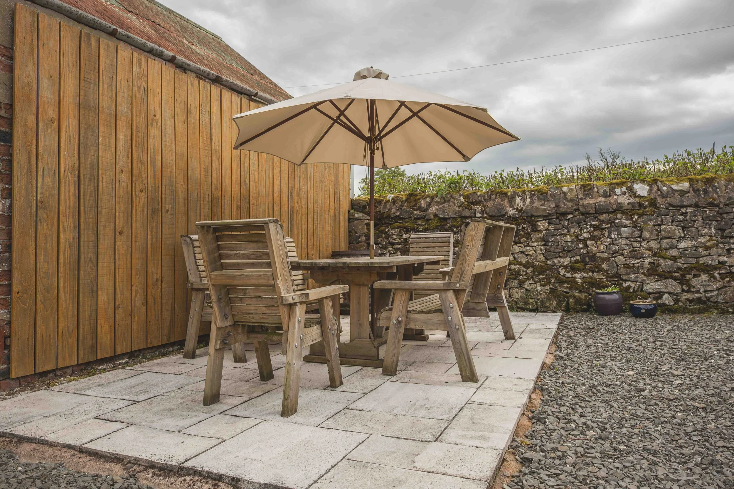 A small outdoor patio area with a wooden table, four wooden chairs, and a large beige patio umbrella. The patio is paved with light-colored stones and is bordered by a stone wall with some green plants growing behind it. There is a wooden fence on the left side, and the sky appears overcast.