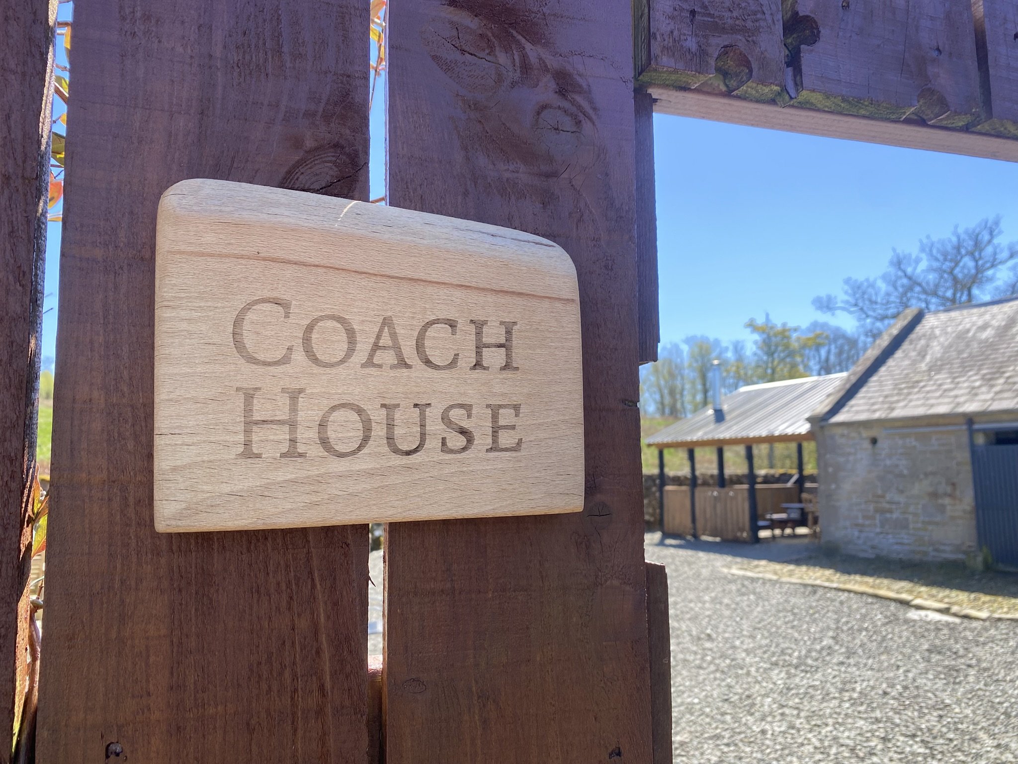 Close-up of a wooden sign with the word 'COACH HOUSE' engraved, attached to a wooden post. In the background, there is a building with a sloped roof and trees under a clear blue sky.