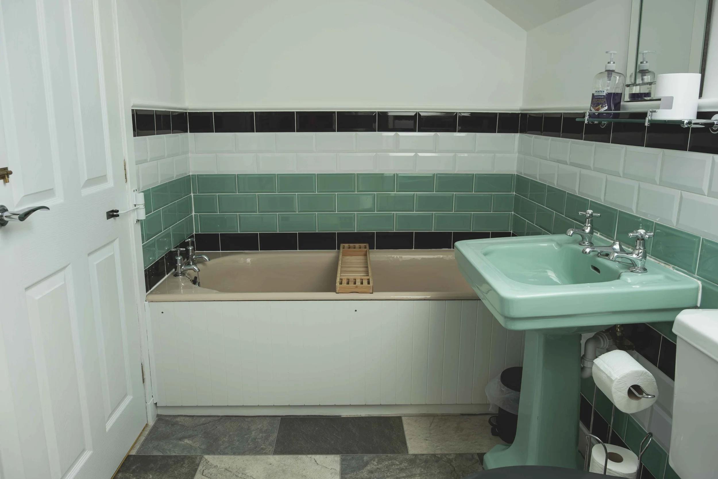 A bathroom with a dark green tiled sink and white tiled walls with black accent tiles. There is a frosted green bathtub with a wooden bath caddy, a white door, a mirror, a shelf with soap and paper towels, a toilet paper roll, and a trash bin.
