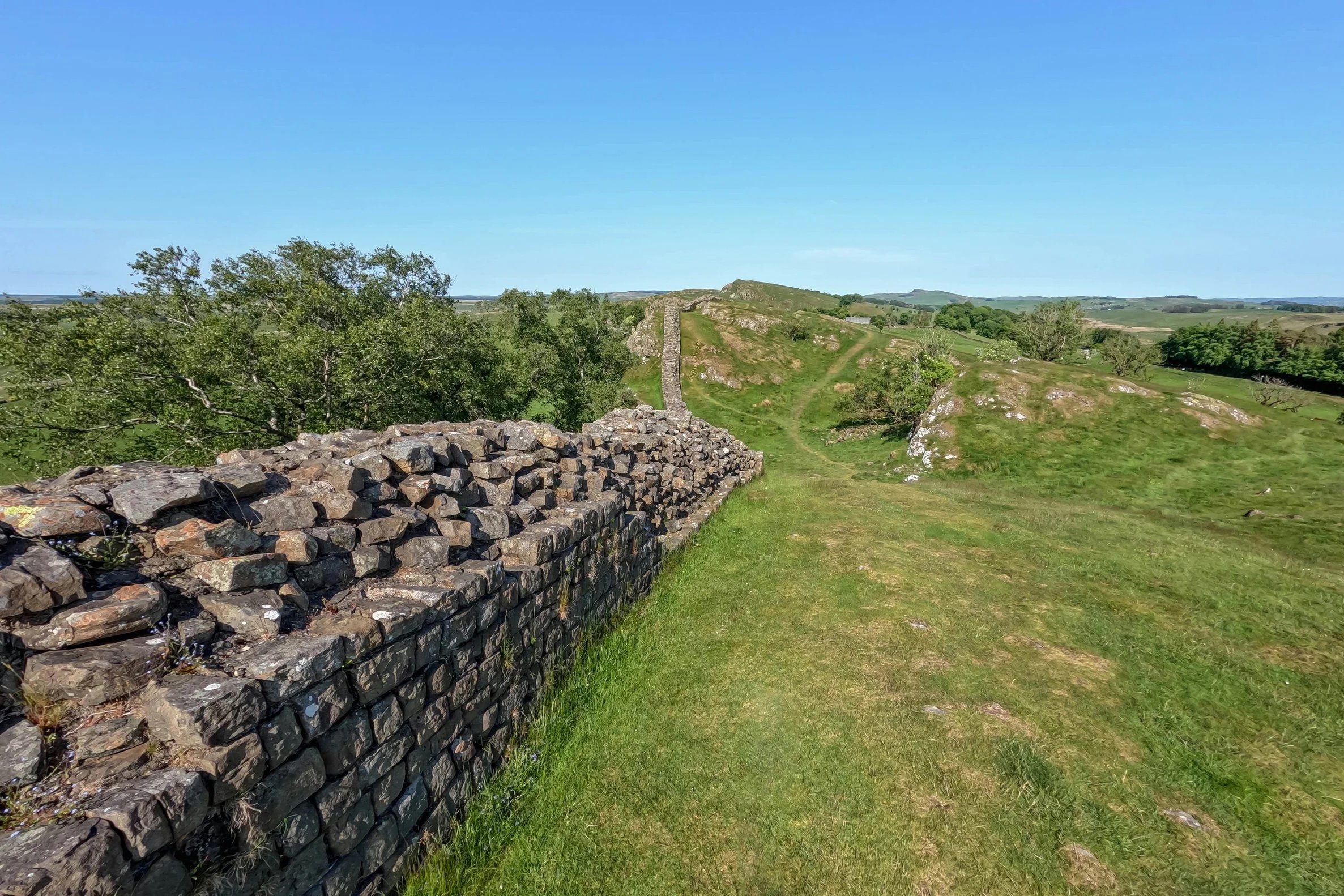 Scenic landscape with stone wall, grassy path, green trees, rolling hills, and a clear blue sky.