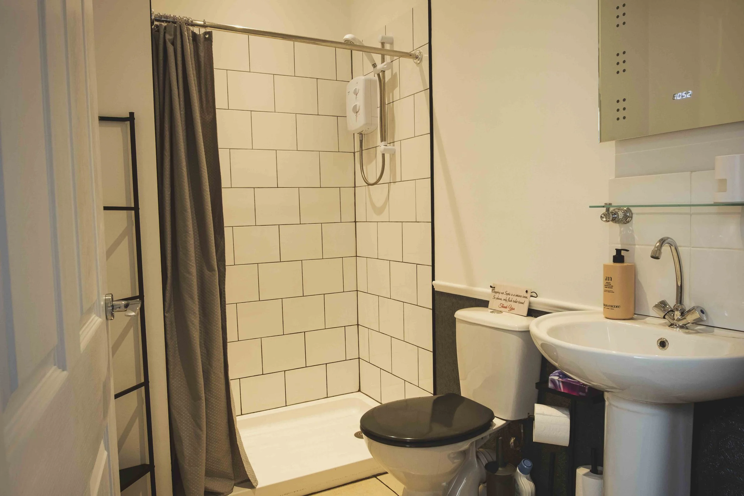 A small bathroom with a walk-in shower, toilet, and sink. The shower has white tiles and a gray curtain, a shower head, and a digital display on the wall. The sink has a round mirror, a soap dispenser, and some toiletries. There is a small black cabi