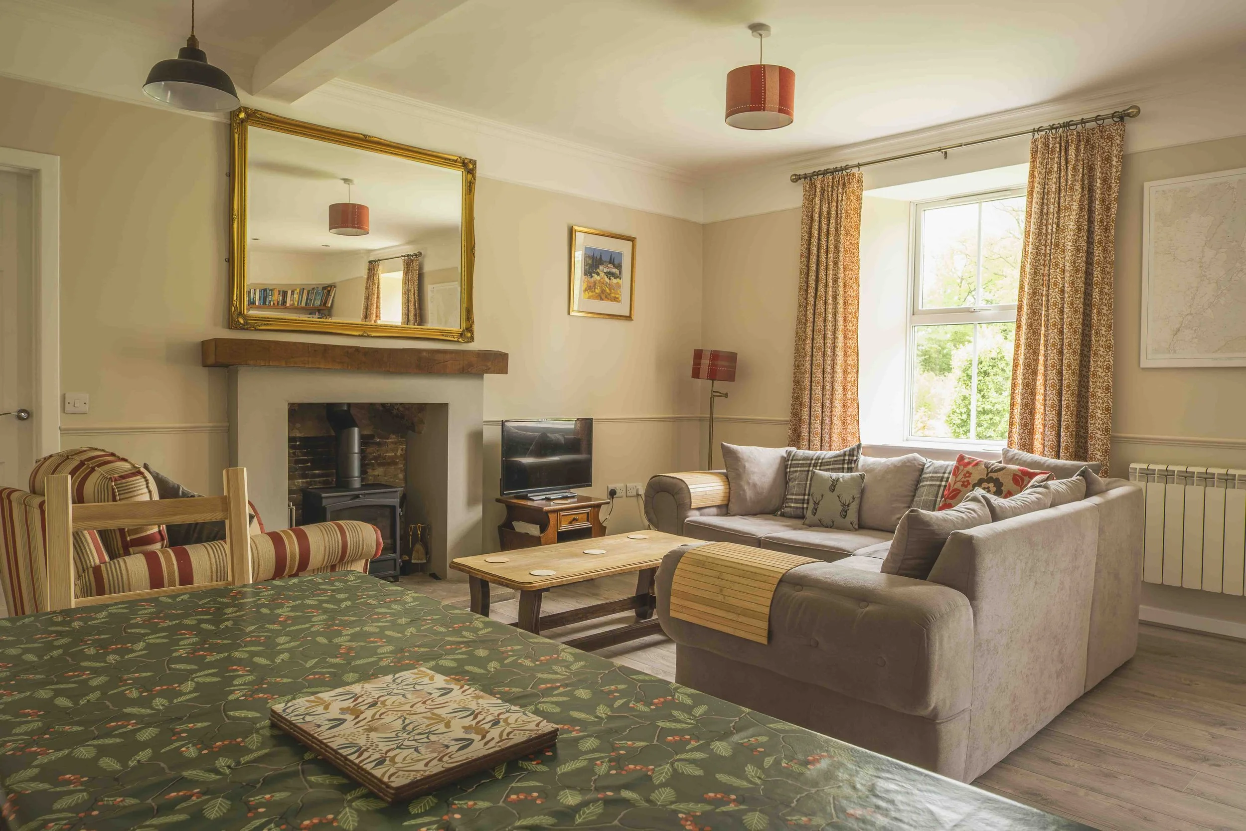 A cozy living room with a large beige sectional sofa, a wooden coffee table, and a patterned armchair. There is a fireplace with a wooden mantel and a large gold-framed mirror above it. The room has a window with floral curtains, a small TV on a wood