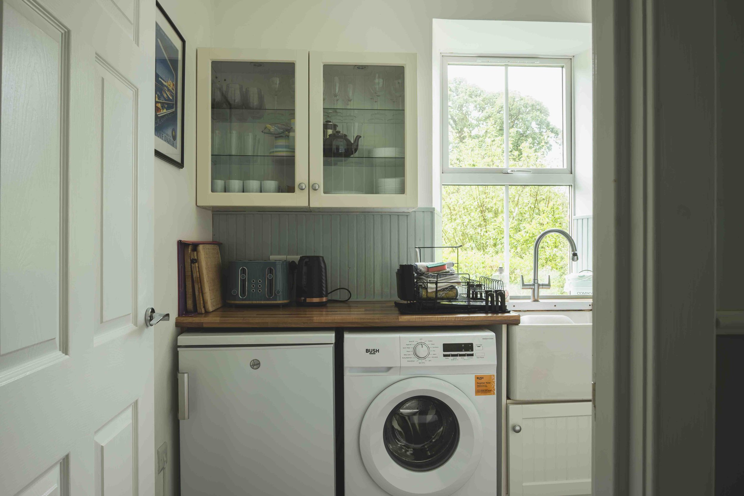A compact laundry room featuring a washing machine beneath a wooden countertop, a wall-mounted cabinet with glass doors holding dishes and glasses, a window showing green trees outside, and various small appliances and items on the countertop.