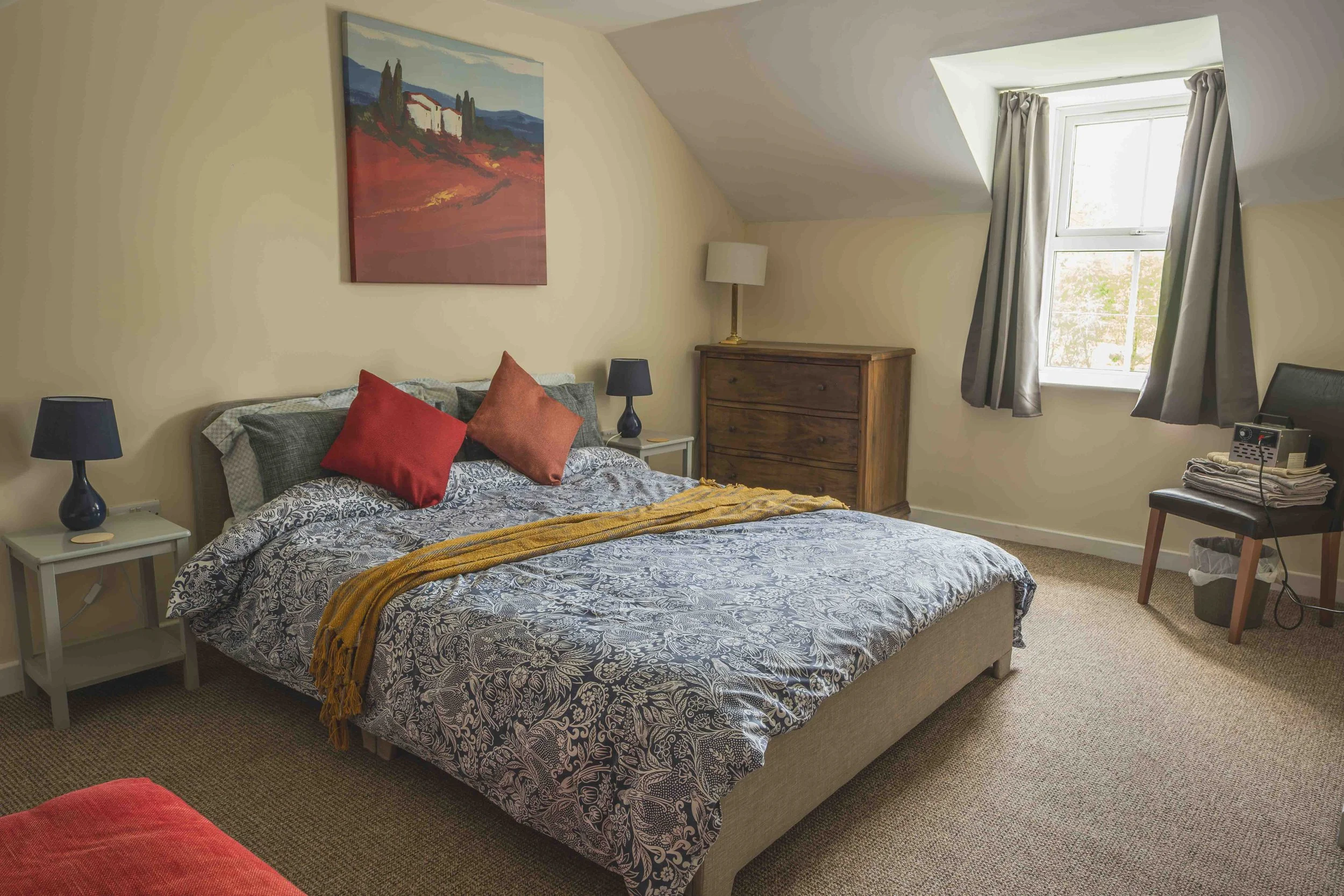 A cozy bedroom with a bed covered in black and white patterned bedding, accented by red and orange throw pillows. Two nightstands with black lamps flank the bed. A wooden dresser with a lamp and a small table with a phone and folded towels are near a