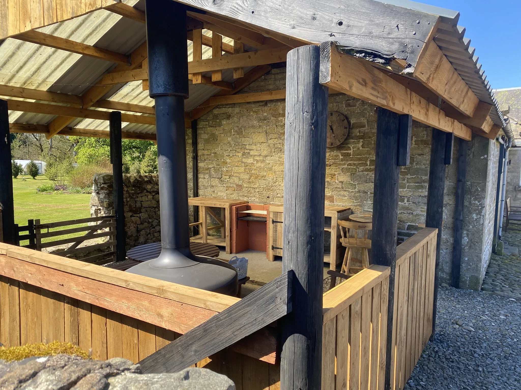 A wooden undercover barbecue are with a corrugated metal roof, supported by black posts and a large wooden post, attached to a stone building. There are trees and a grassy area in the background.