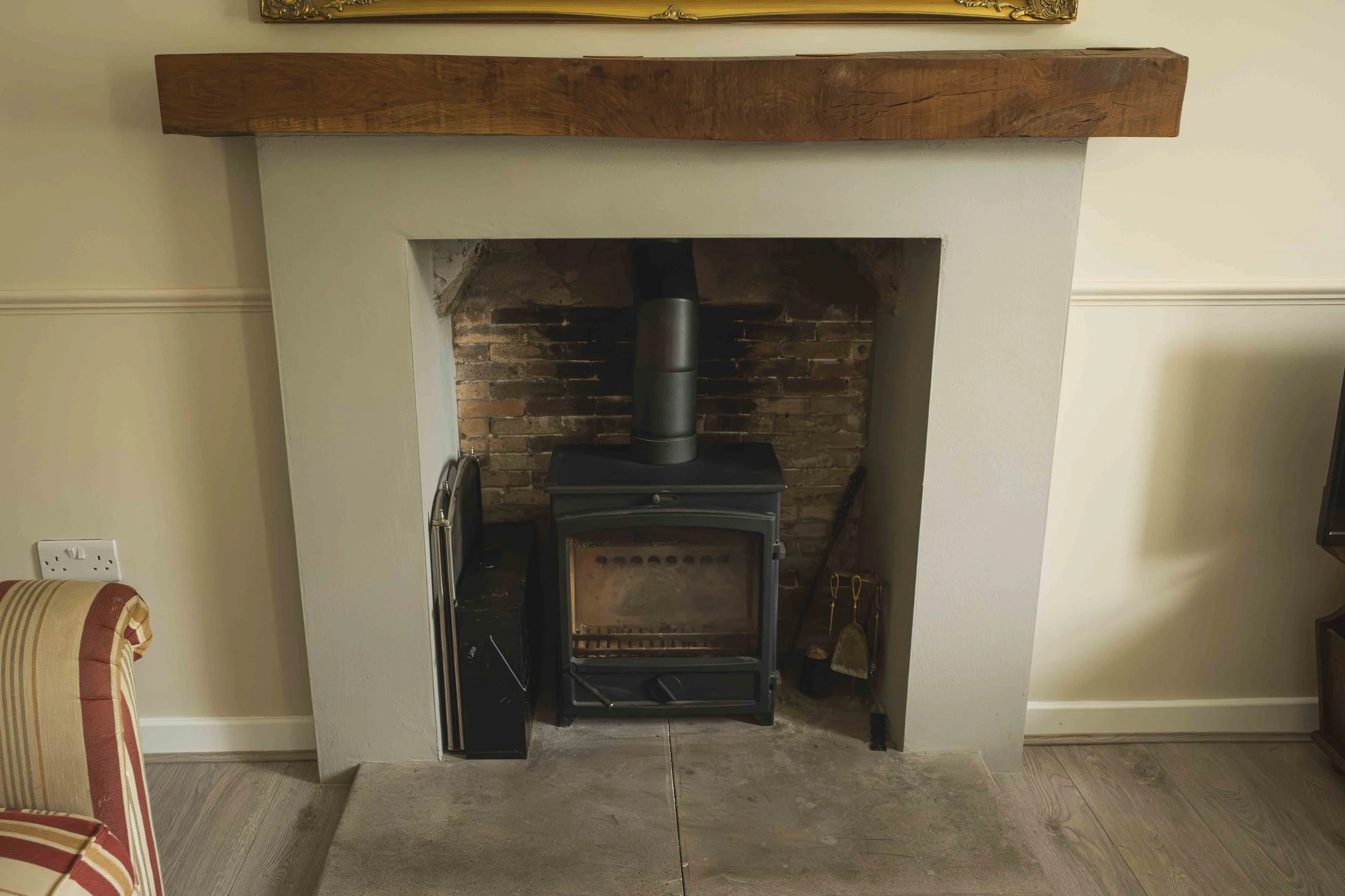 A living room fireplace with a wooden mantel and a brick interior, containing a black wood stove. To the left, part of a striped upholstered chair is visible. The wall behind is light-colored with a socket near the floor, and a framed picture is part