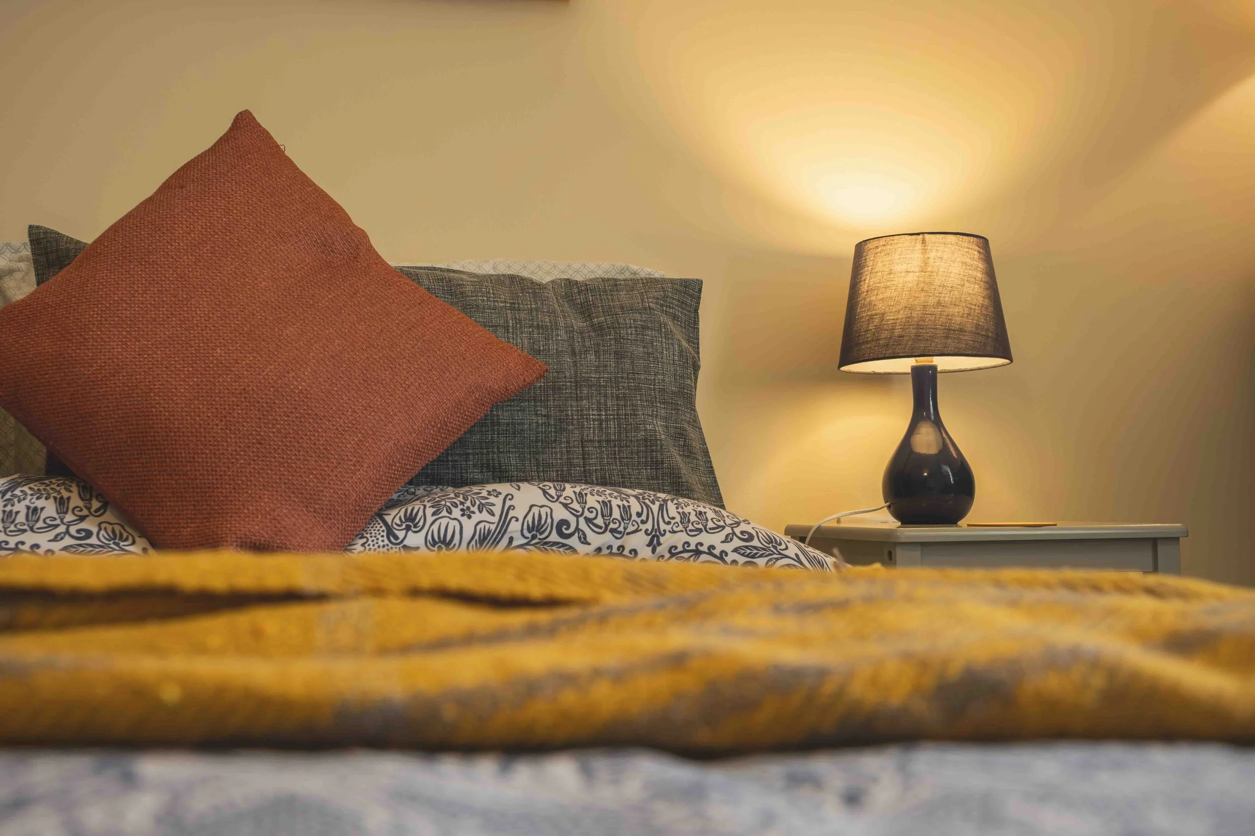 Close-up of bed linens with a patterned bedsheet, a grey pillow, a rust-colored throw pillow, and a bedside lamp on a nightstand, taken from a low angle.