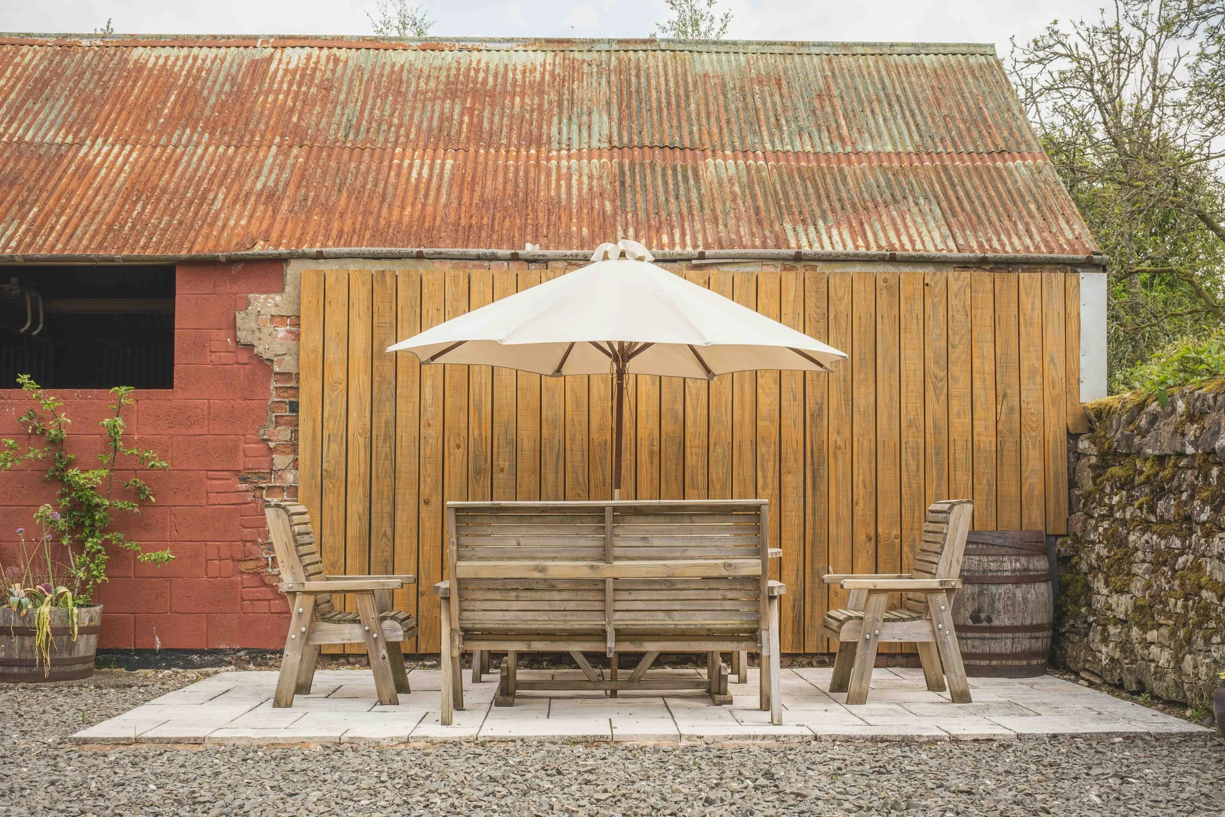 A rustic outdoor patio area with a wooden table and benches, a large white patio umbrella, and a brick and wooden horse fencing in front of a weathered building with a corrugated metal roof.