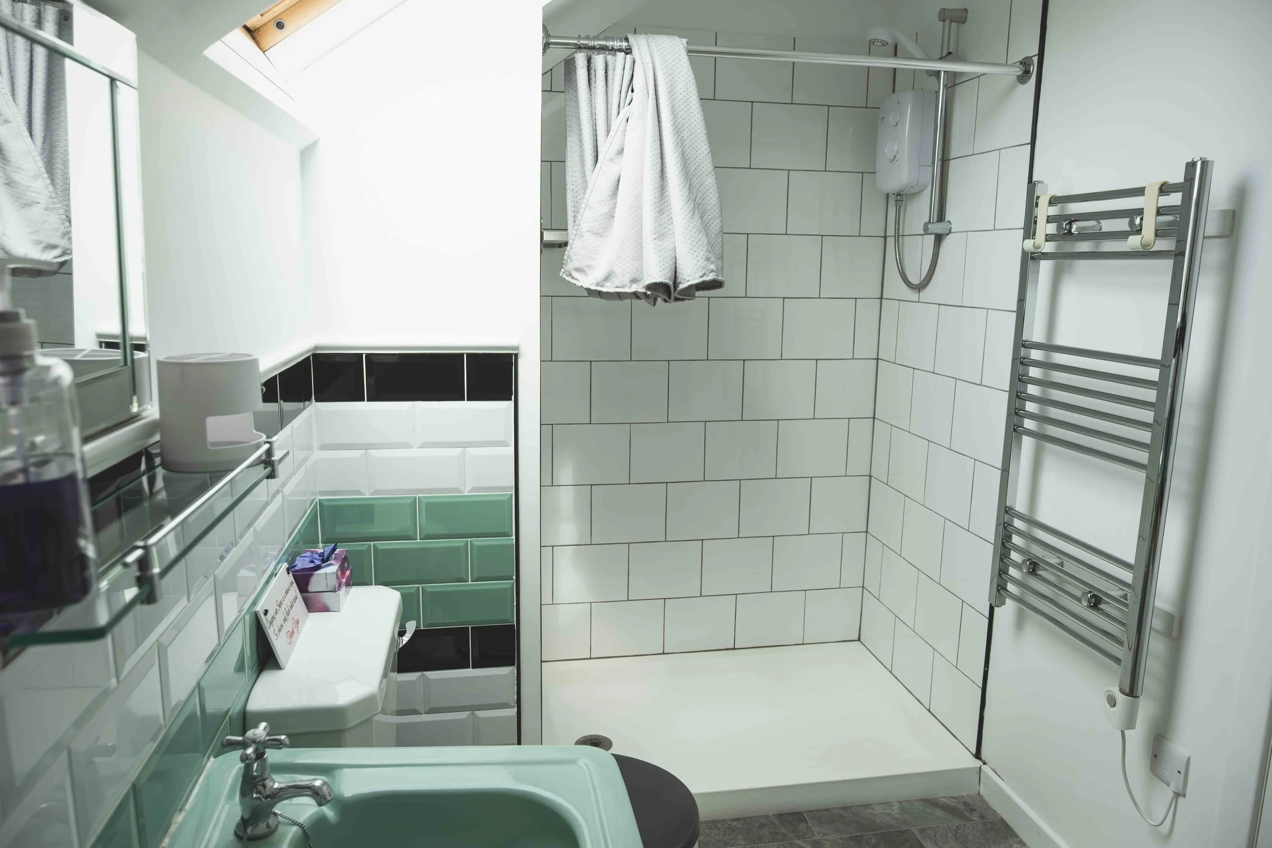A modern bathroom with white tiled walls and a skylight window. It features a green sink, a towel rack, a shower area with a curtain, and toiletries on a shelf.