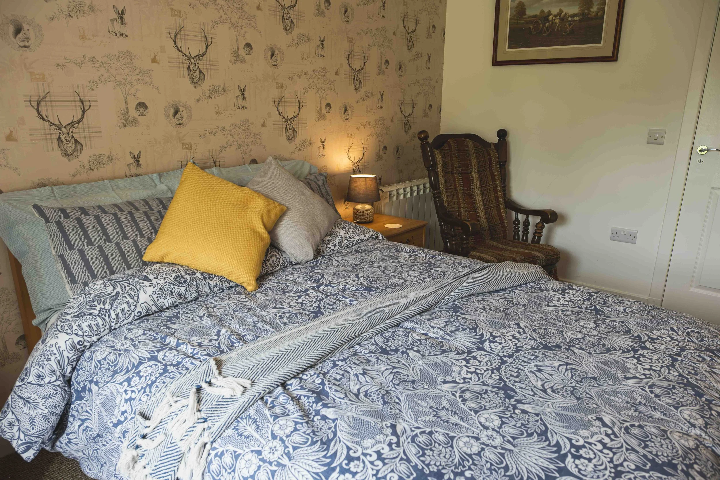 A bedroom with a bed covered in a blue and white floral patterned duvet and multiple pillows, a bedside table with a lampshade, a wooden chair with a checkered cushion, and wall decorations including framed pictures and wallpaper with animal motifs.