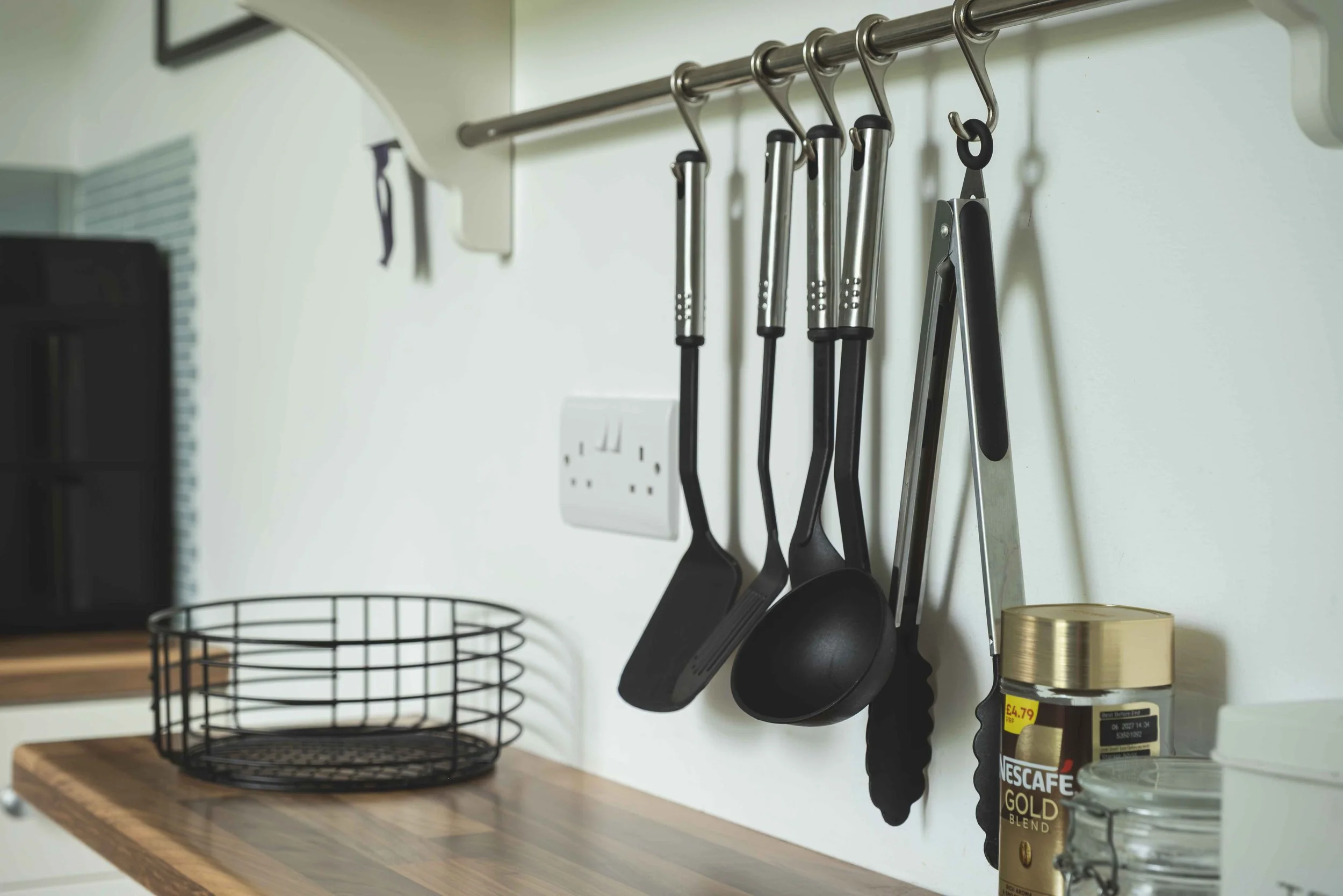 Kitchen countertop with a black wire basket, a set of hanging black kitchen utensils, a Nescafe Gold coffee jar, and a small glass jar.