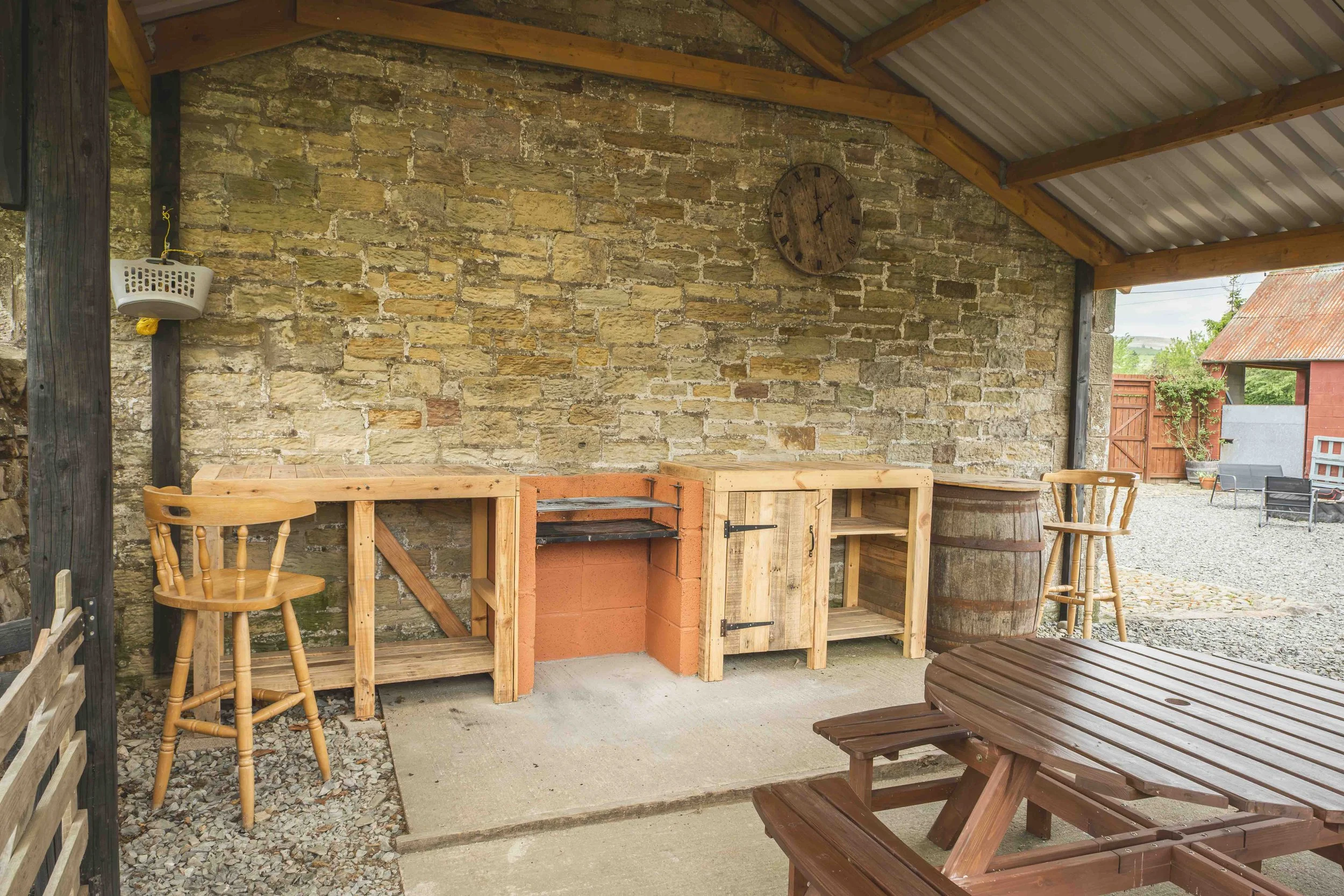 A rustic outdoor patio area with wooden furniture, including a table, chairs, and a bar, against a stone wall with a large round clock. There is a wooden roof overhead and a gravel and concrete ground, with outdoor scenery visible in the background.
