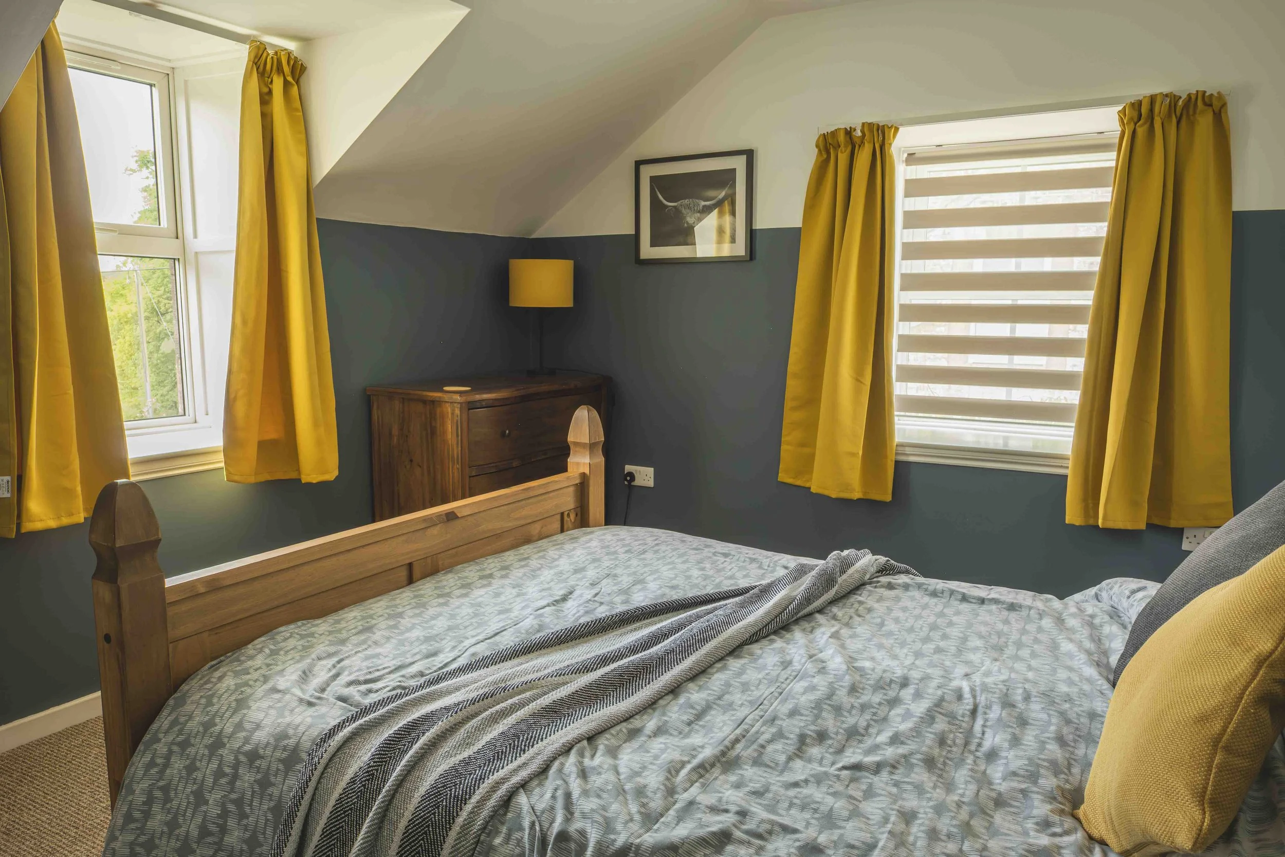 A cozy bedroom with a wooden bed, green and yellow accents, and two windows with yellow curtains letting in natural light.