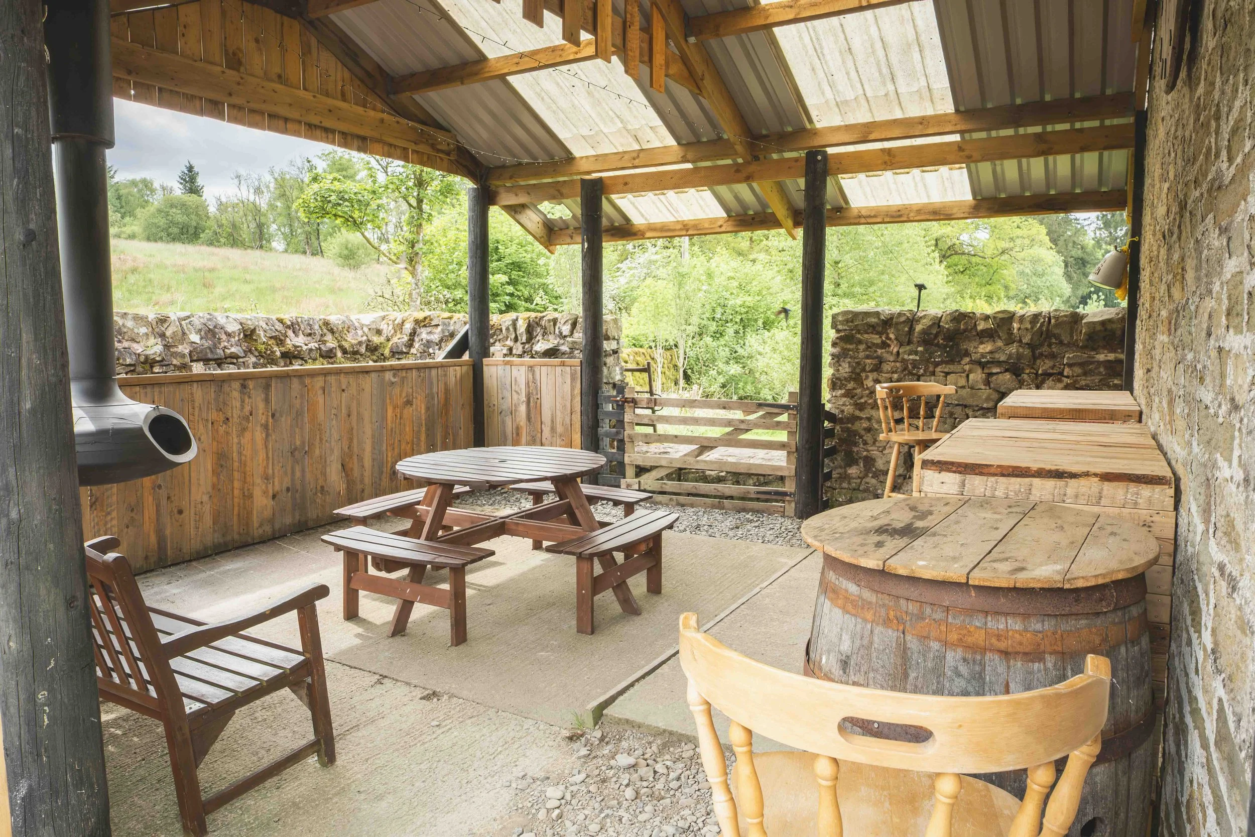 Covered outdoor patio with wooden picnic tables, chairs, a stone wall, and a barrel table, overlooking green trees and a grassy hill.