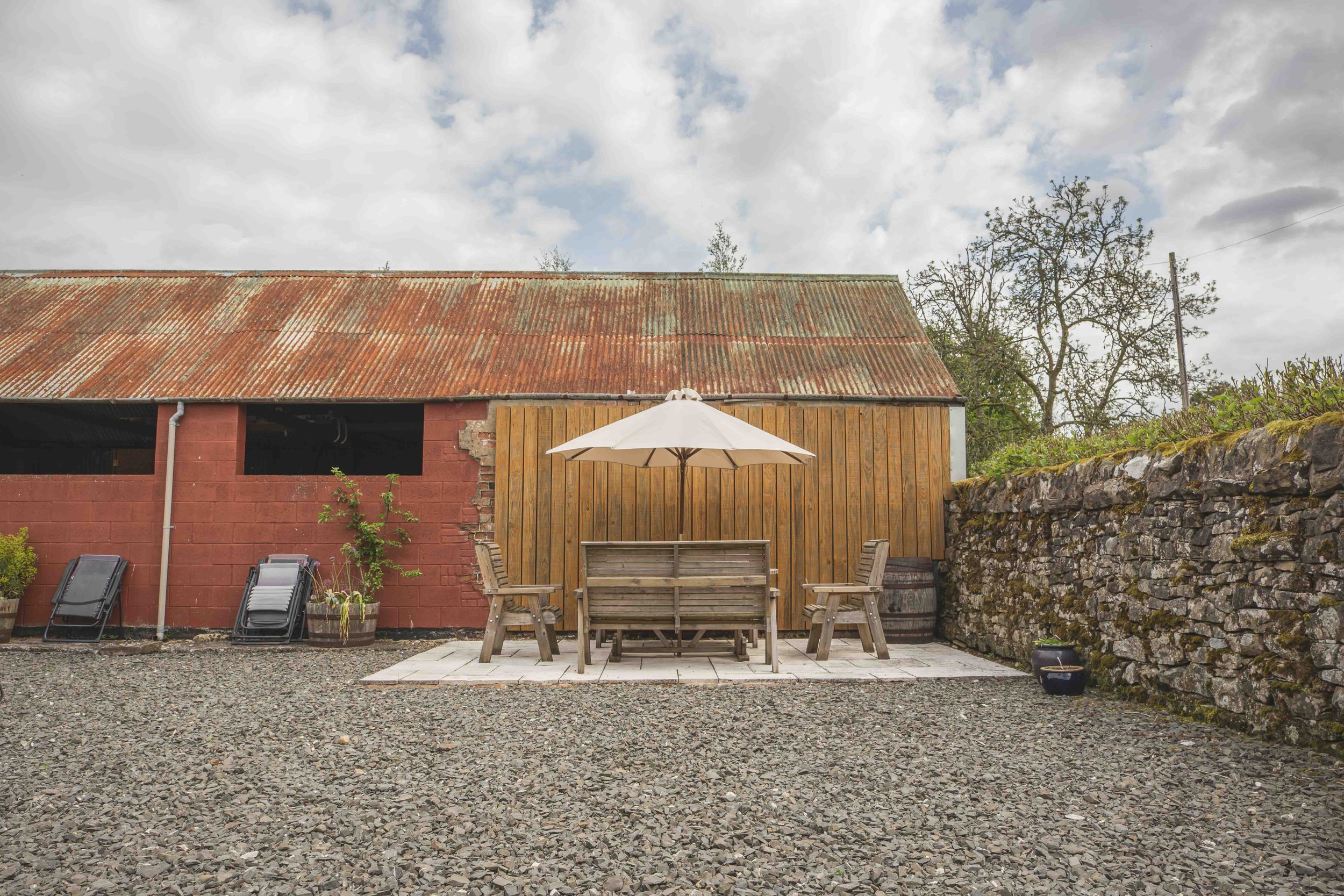 A rustic backyard scene features a weathered red barn with a corrugated metal roof, a wooden patio area with outdoor furniture, a white patio umbrella, and a stone wall on the right. There are two lounge chairs leaning against the barn, and the groun