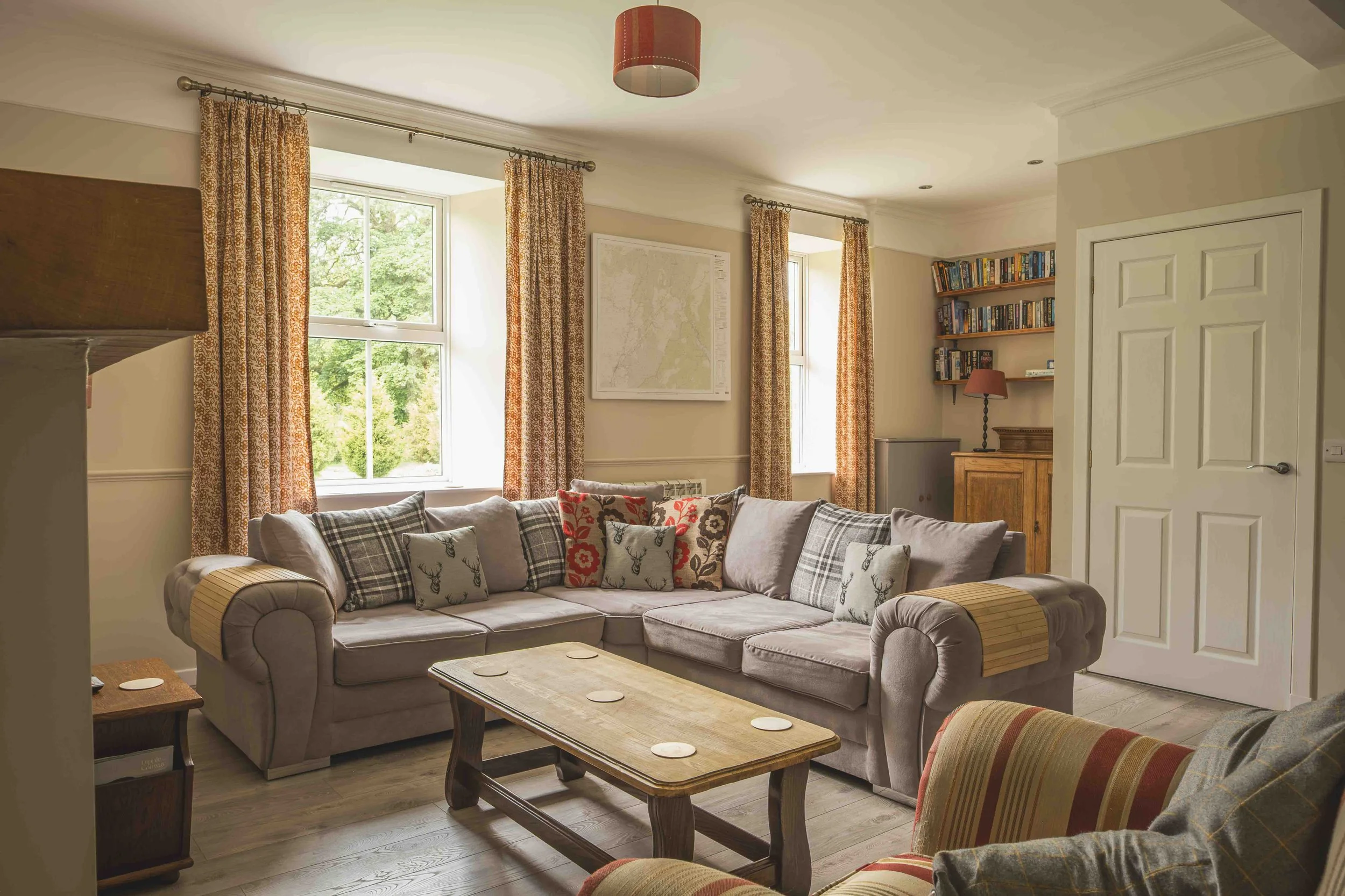 Living room with large windows covered by patterned curtains, a plaid couch with decorative pillows, a wooden coffee table, a bookshelf filled with books, a small table lamp, and a map on the wall.