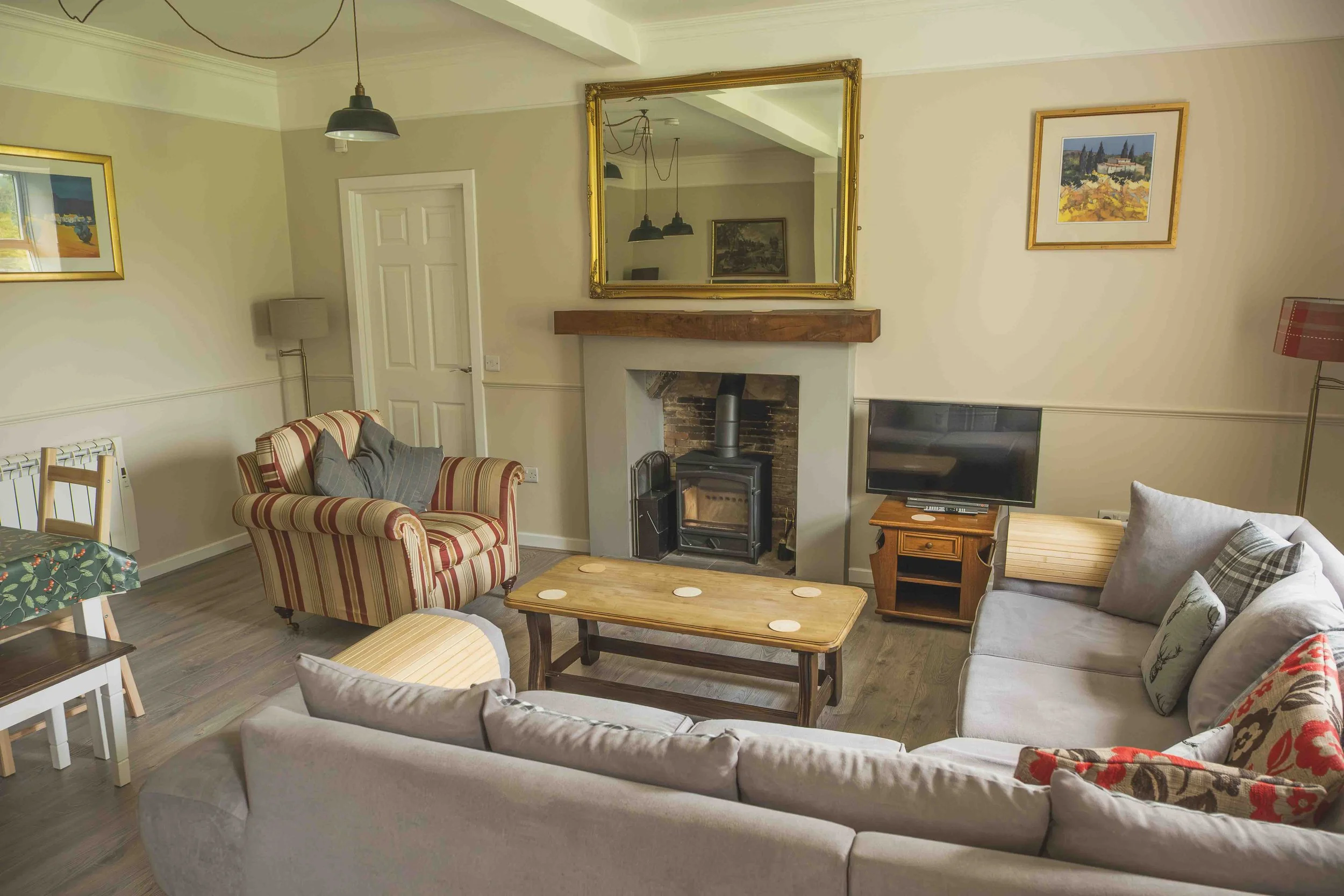 Living room with cream-colored sofas, a wooden coffee table with coasters, a fireplace with a wood stove, a TV on a small wooden stand, framed artwork on the walls, and a large mirror above the fireplace reflecting the room's ceiling and lighting fixtures.