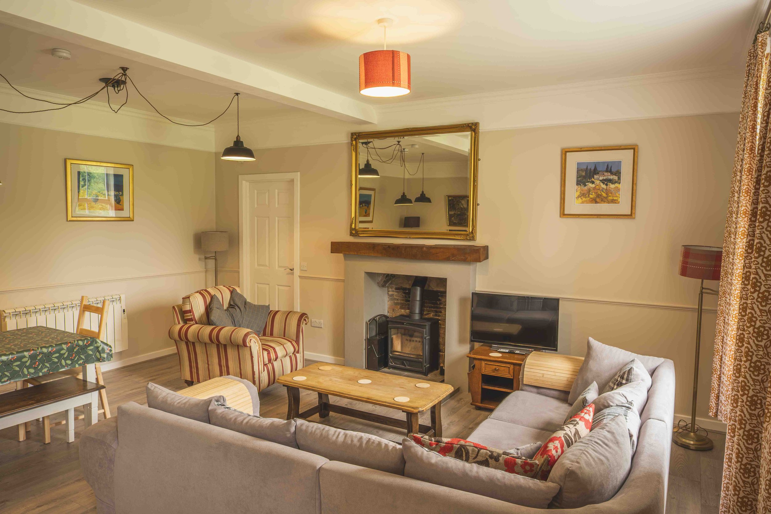 Living room with beige walls, a white ceiling, and wooden flooring. Contains a gray upholstered sofa with patterned cushions, a wooden coffee table, a flat-screen TV, a fireplace with a wooden mantel, and wall art paintings. There are two sets of cur