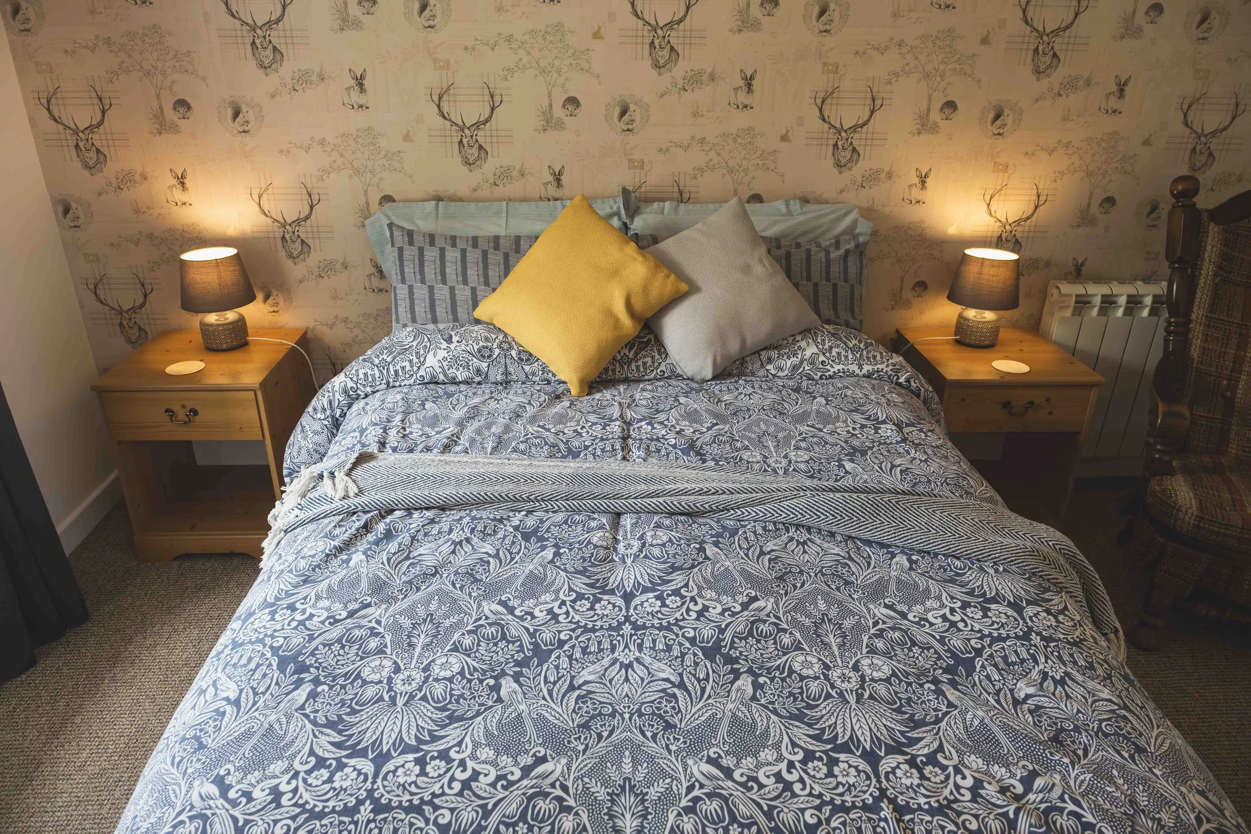 A cozy bedroom with a bed covered in a blue and white floral patterned comforter, decorated with three pillows: a yellow one and two neutral-colored ones. There are two matching bedside tables, each with a black lampshade and a small decorative item,