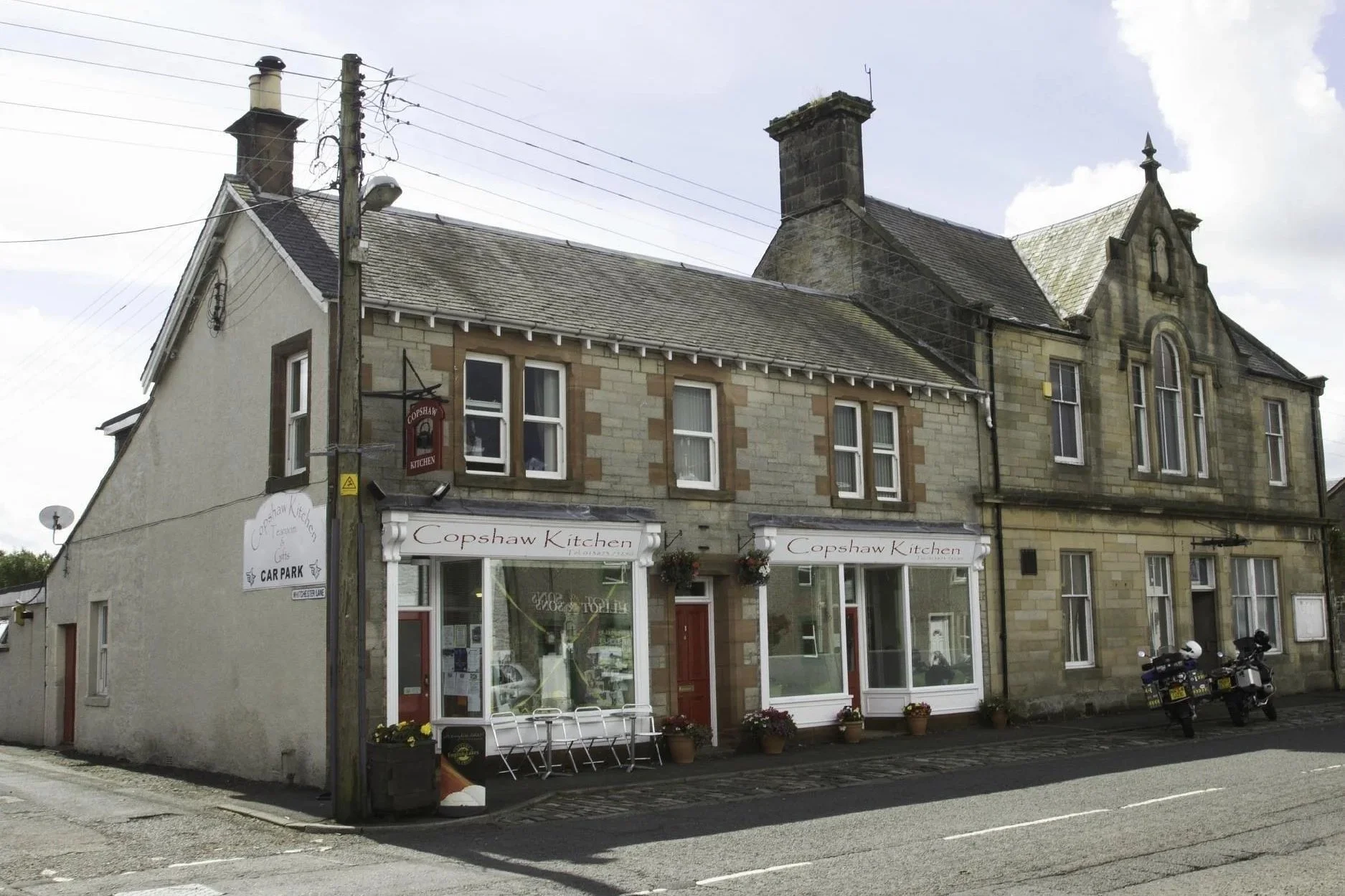 A two-story stone building with a cafe called Coshaw Kitchen on the ground floor. The building has multiple windows, a gabled roof, and a chimney. There are potted plants and hanging baskets outside the cafe, with a sign indicating a car park. A motorcycle is parked in front of the building.