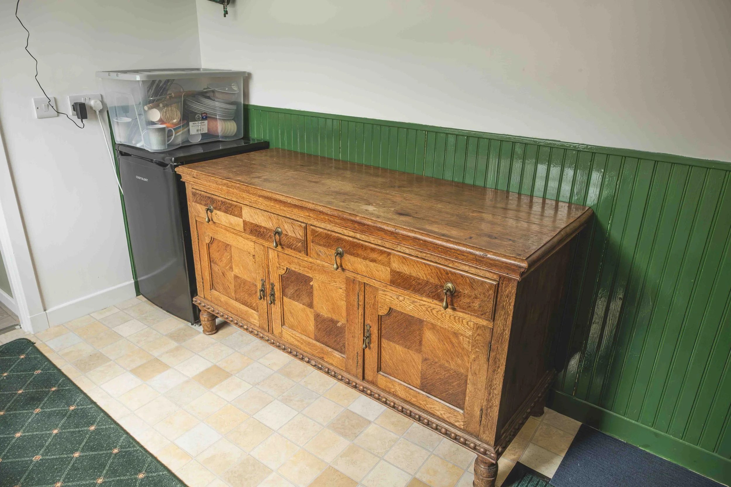 A wooden sideboard with drawers and cabinets next to a green paneled wall, with a plastic storage bin on top containing kitchen utensils and cups, in a room with tiled flooring.