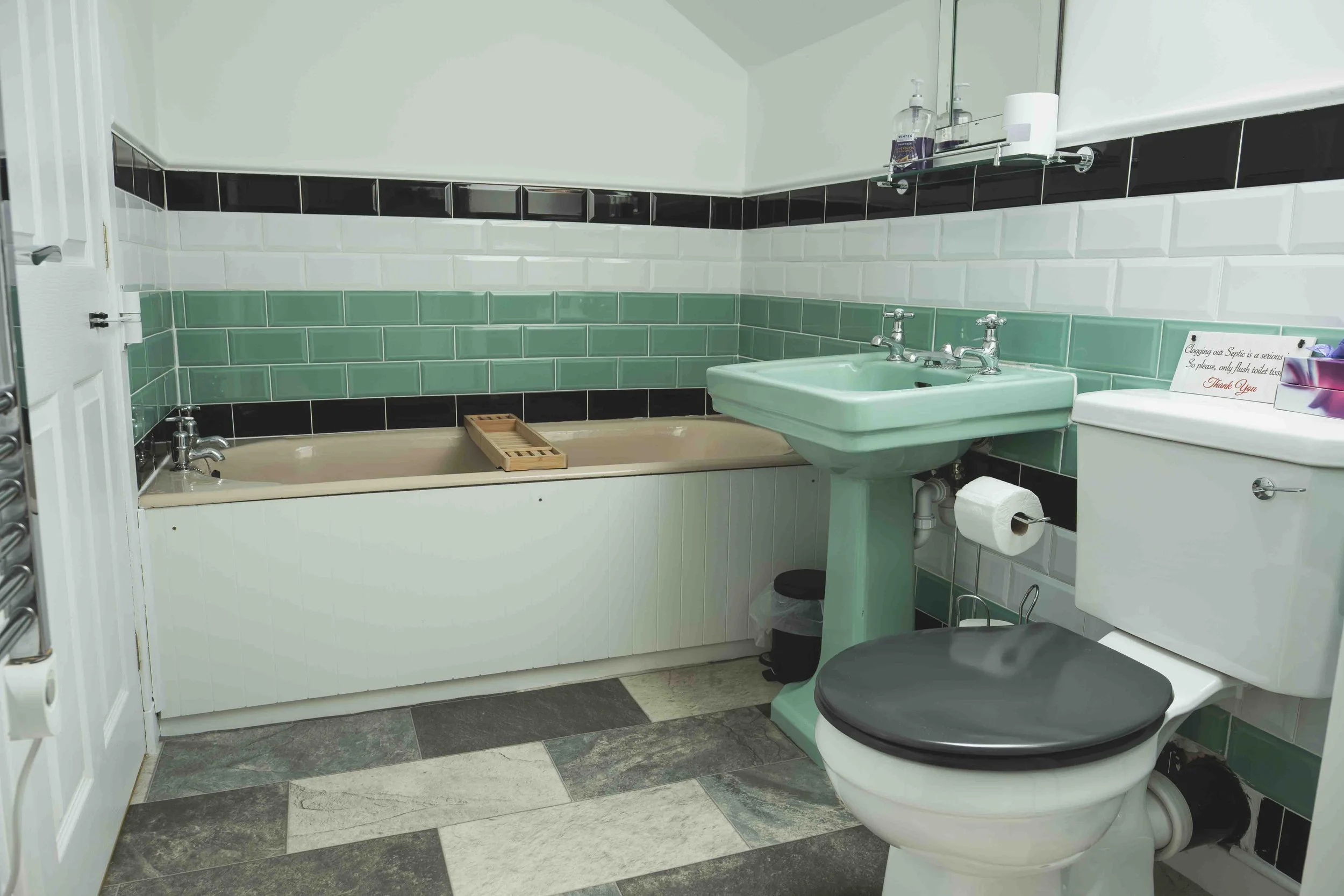 A bathroom with subway tile walls in green, white, and black, a bathtub with a wooden bath tray, a mint green sink, a toilet with a paper holder, and a shelf with toiletries.