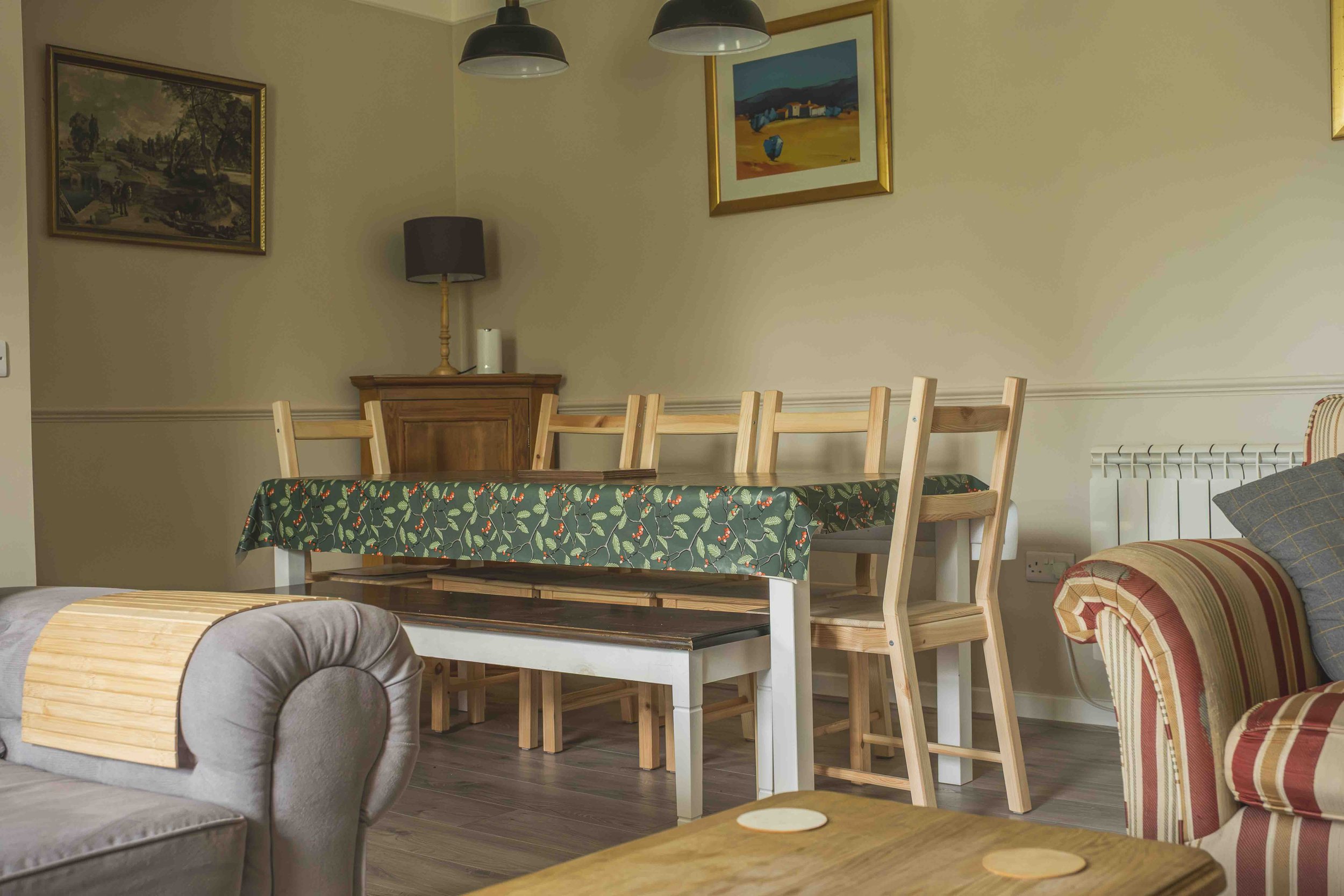 Living room with wooden dining table covered with a green tablecloth with a holly pattern, surrounded by wooden chairs. There is a wooden cabinet with a black lamp, a white candle, and a paper towel roll on top. The room has beige walls with framed p