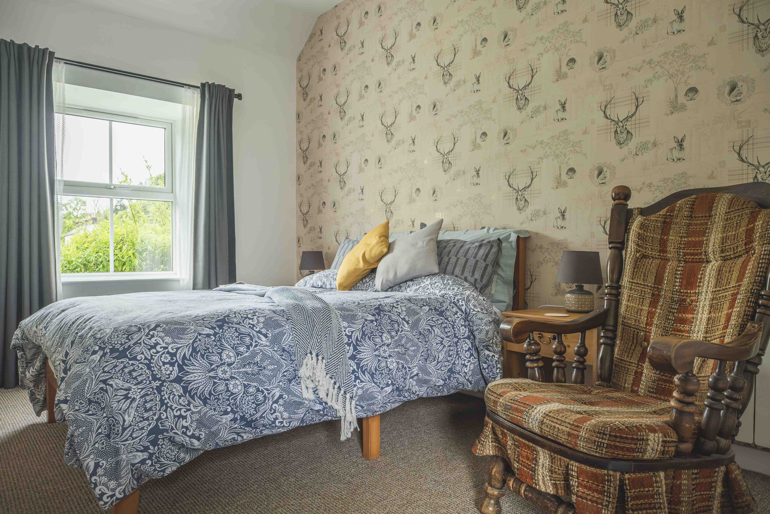 A bedroom with a bed covered in a black and white floral patterned bedspread, several pillows including a yellow one, and a window with natural light and greenery outside. The wall behind the bed features wallpaper with images of deer heads, rabbits,