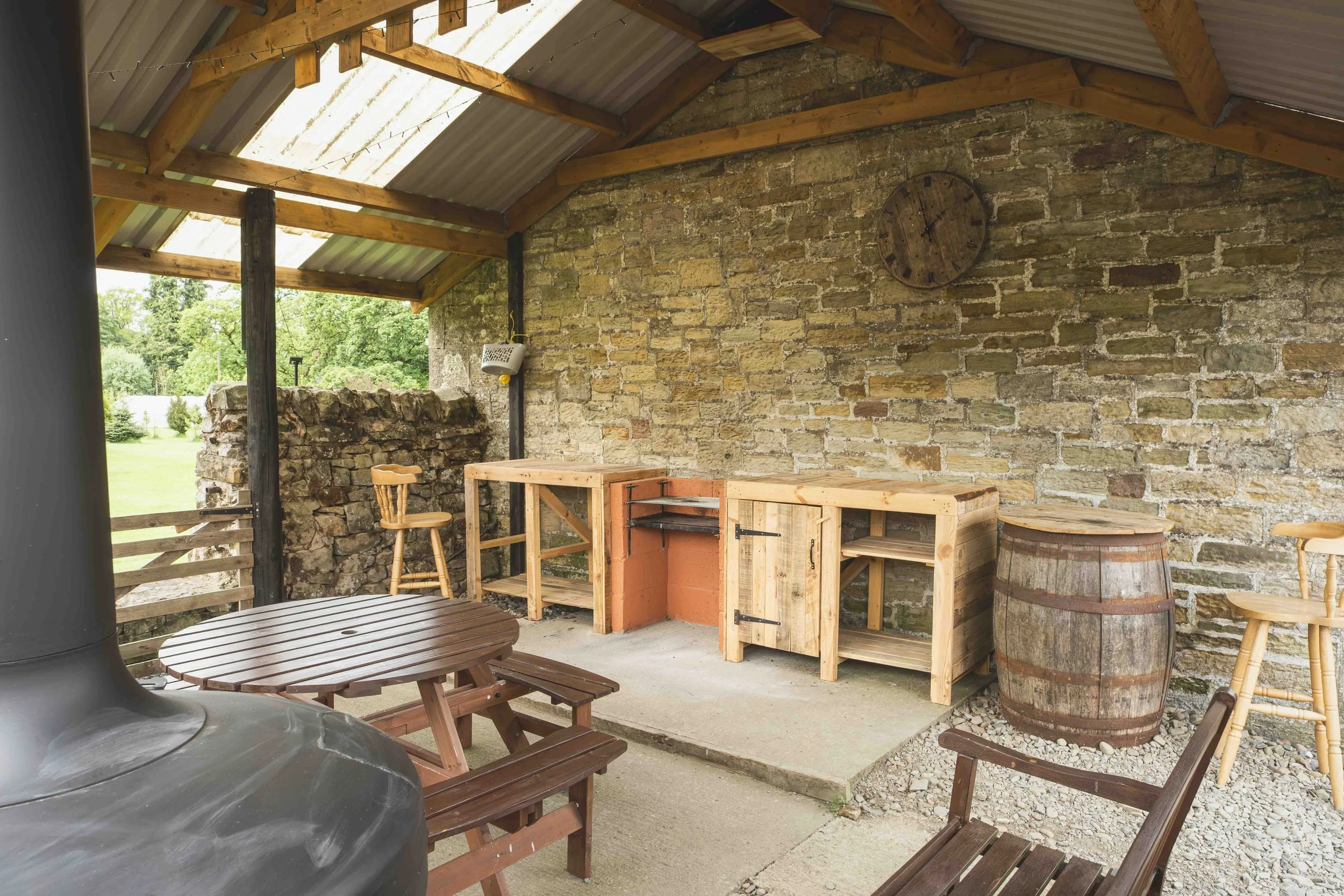 An outdoor patio area with a wooden ceiling, brick wall, and various outdoor furniture including a round wooden table, chairs, and a barrel. There is a vintage clock on the brick wall and a stone counter or grill. Green trees are visible outside thro