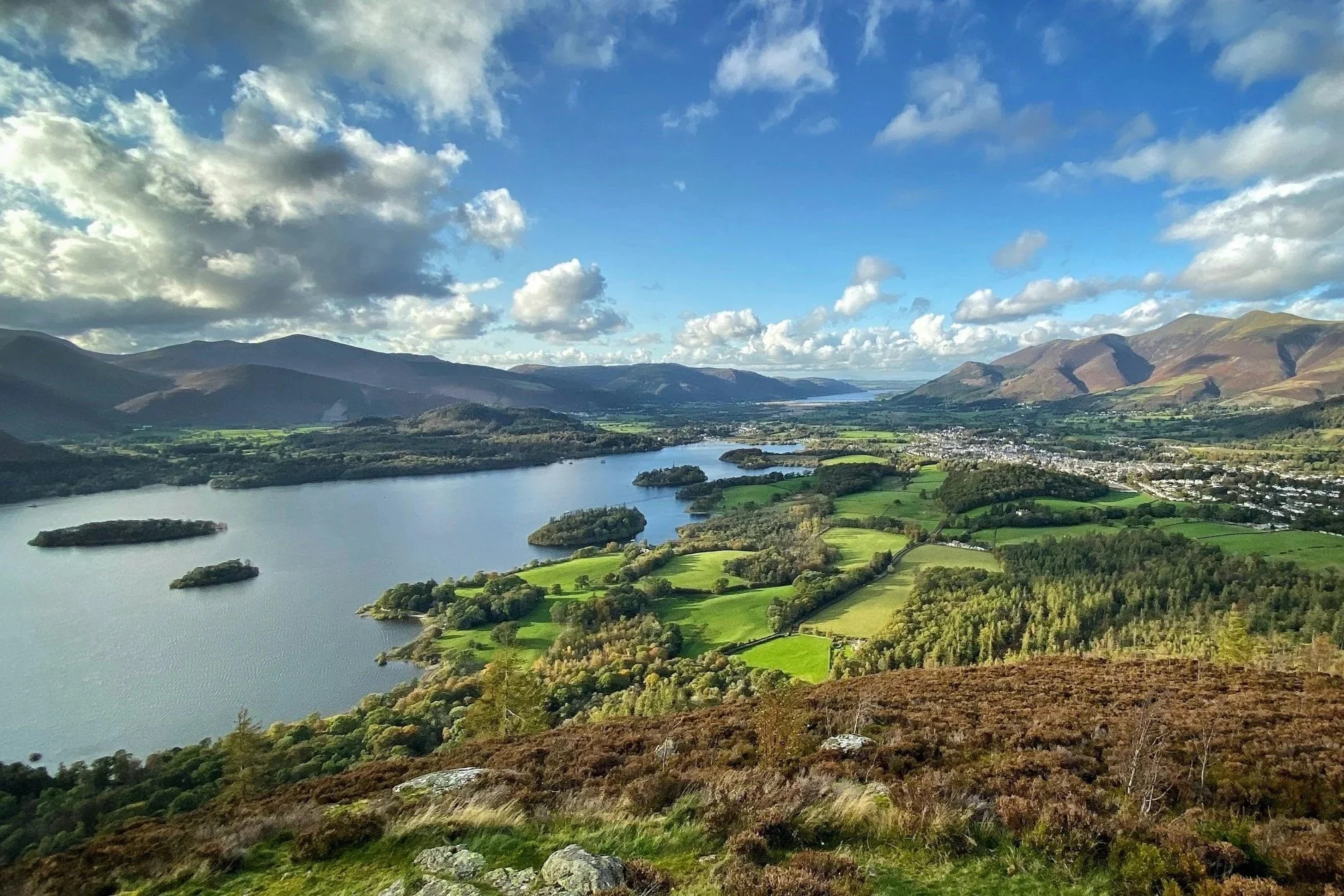 A scenic landscape of a river winding through lush green fields, surrounded by rolling mountains under a partly cloudy blue sky.