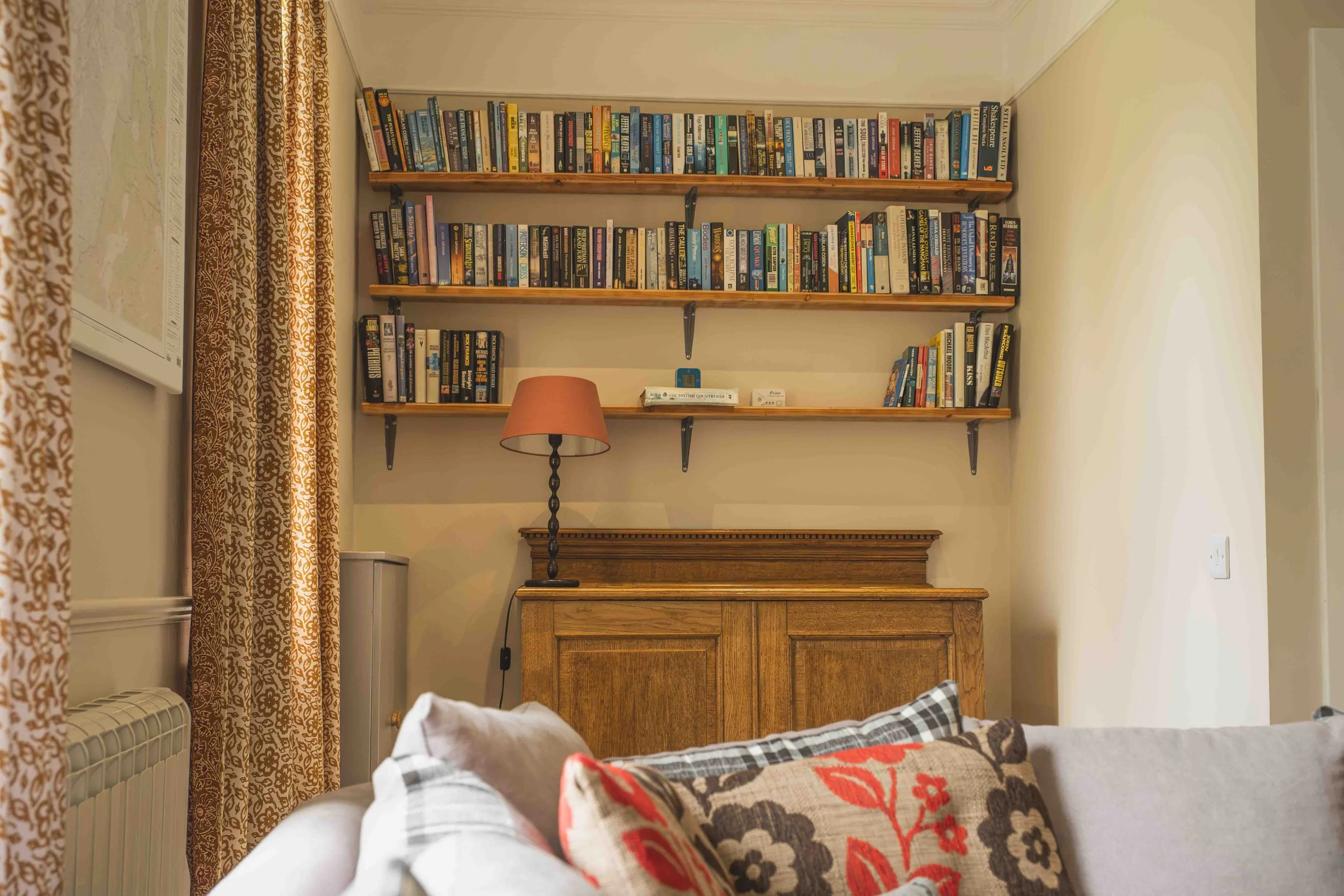 A bedroom corner with a bed, decorative pillows, wooden headboard, bookcase with books, a table lamp, beige curtains with a floral pattern, and a radiator.